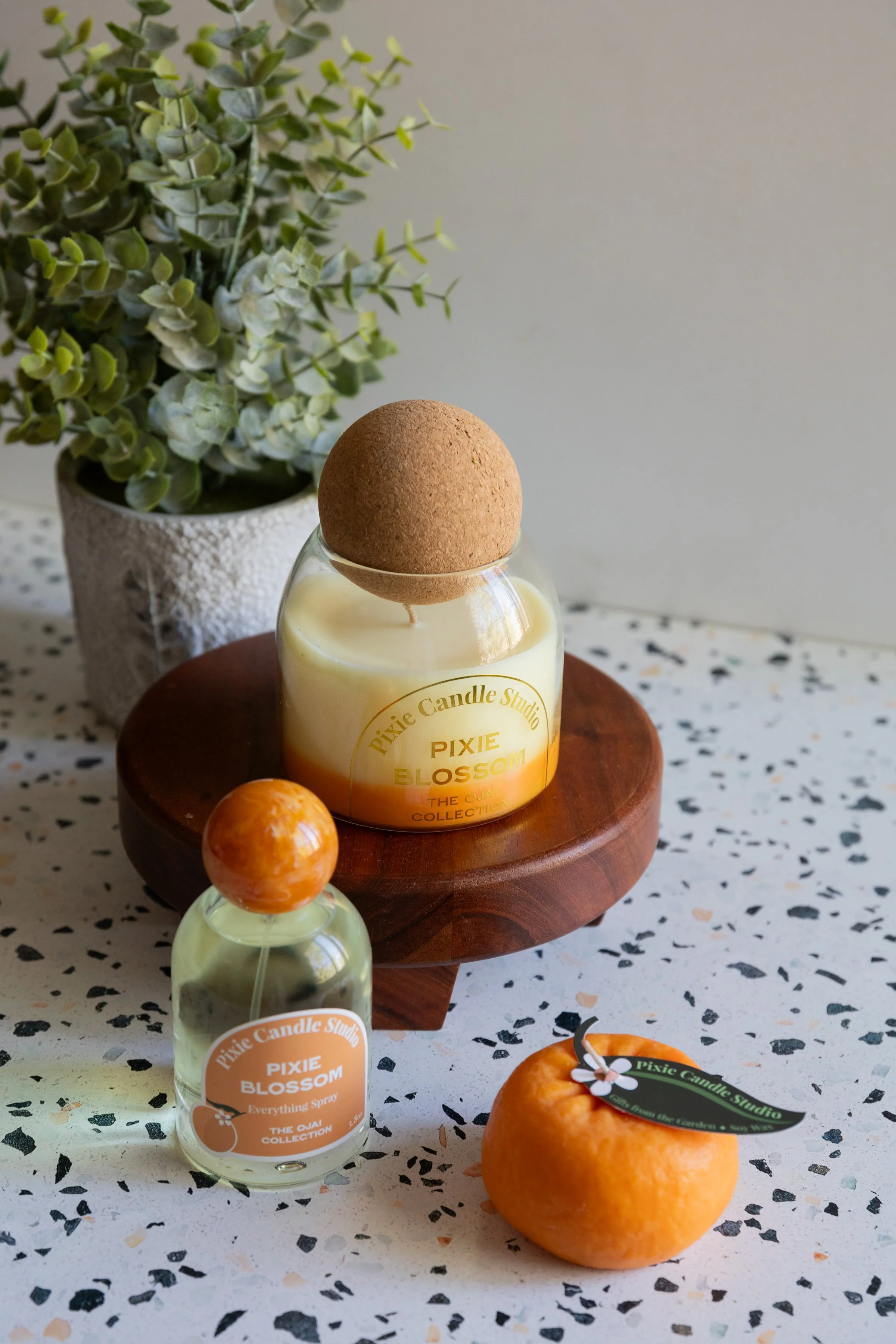 A decorative scene featuring a potted plant with green and white leaves, a wooden tray holding a wax candle labeled 'Pixie Blossom', a small bottle of fragrance spray, and a ripe orange fruit on a speckled white surface.