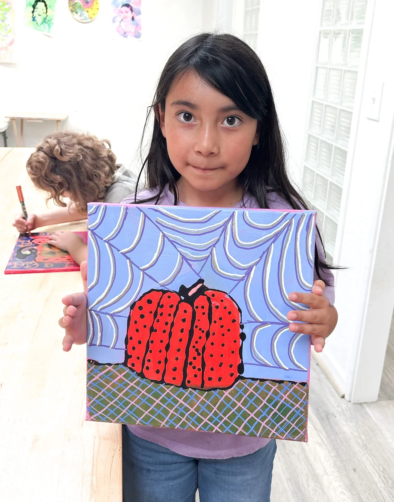 Girl holding a colorful painting of a red pumpkin with black spots and a web in the background.