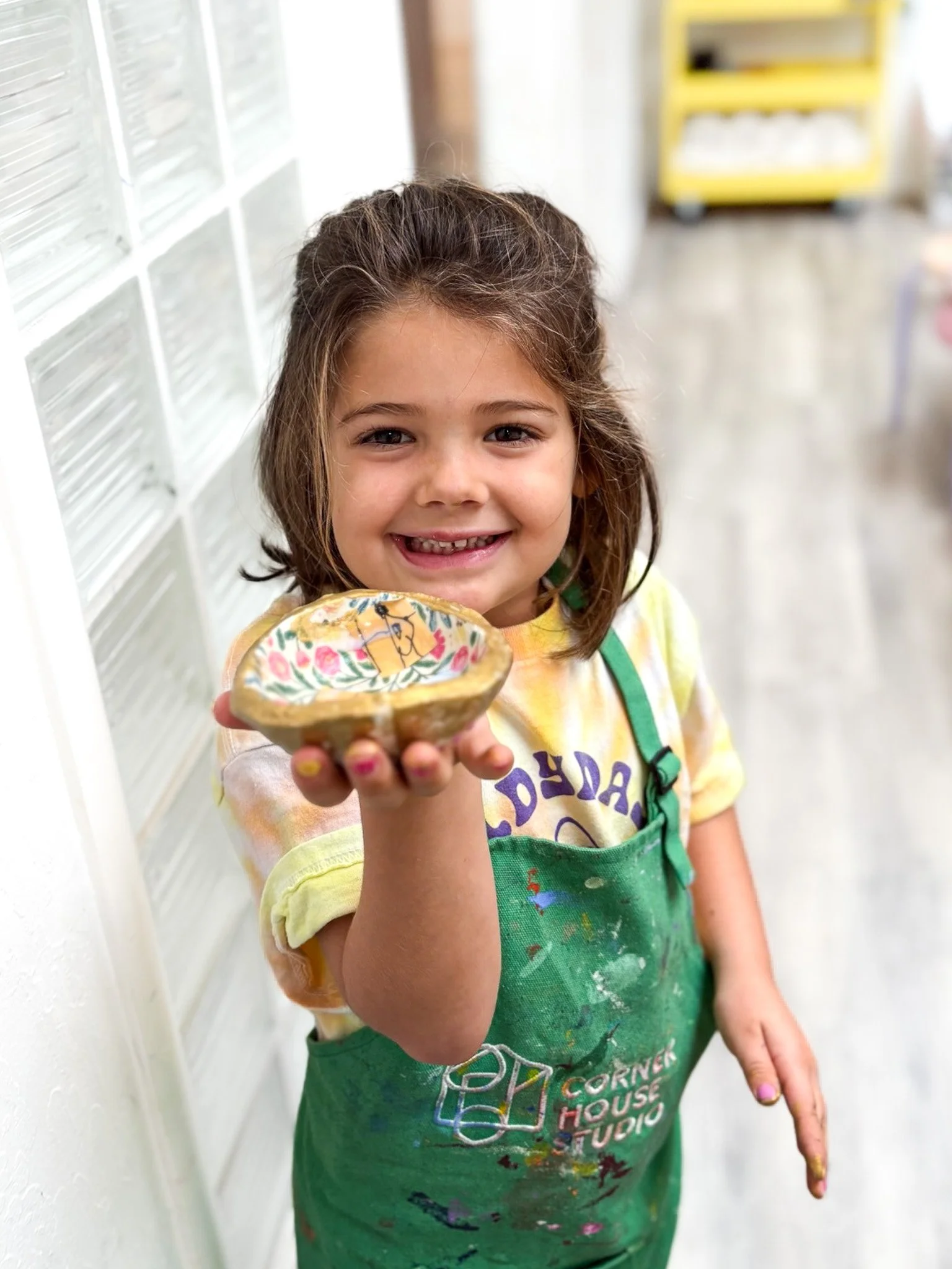 A young girl with brown hair, smiling and showing her teeth, holds a decorated ceramic bowl towards the camera. She is wearing a yellow tie-dye t-shirt and a green apron with paint stains, standing indoors.