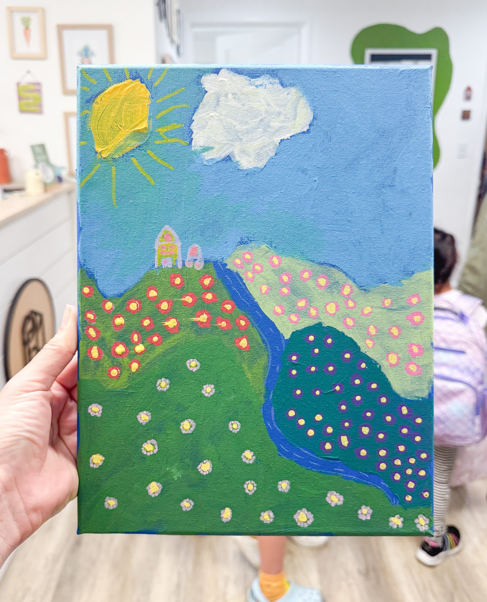 Child's colorful landscape painting depicting a sunny sky with a sun and cloud, a green hillside with pink and yellow flowers, a waterfall, and some houses on a hill.