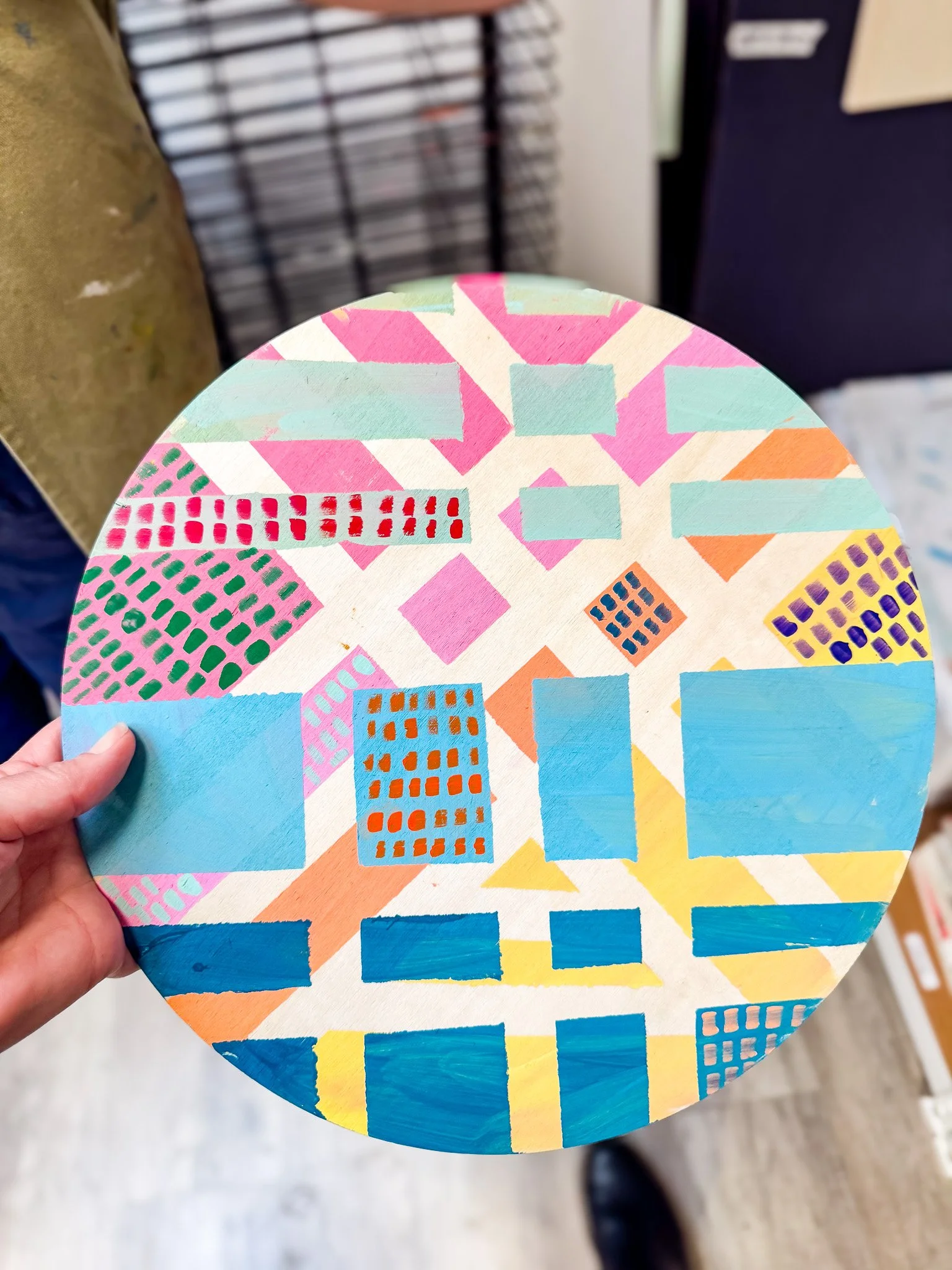 A round canvas with an abstract geometric pattern painted on it, featuring various shapes such as squares, rectangles, and diamonds in bright colors including pink, blue, yellow, orange, purple, and red.