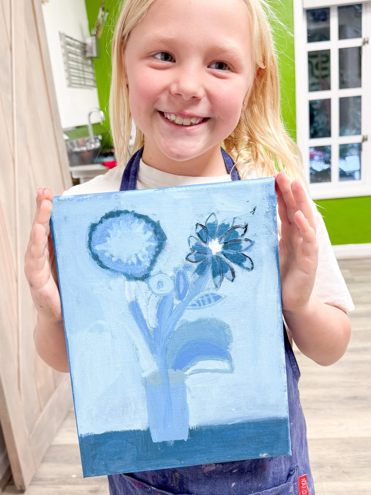 A young girl with blonde hair holding up a painted canvas of blue flowers in a vase, smiling. She is in a room with green walls and a window in the background.