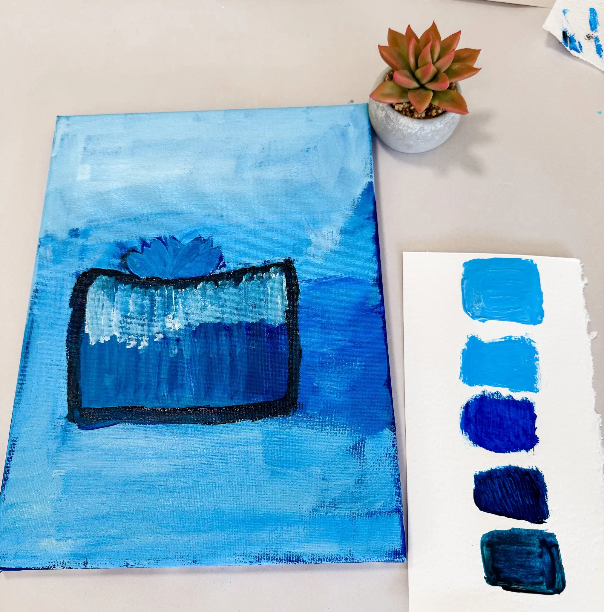 A small canvas painting of a blue succulent in a pot, with a piece of paper showing four shades of blue paint, next to a potted succulent plant.