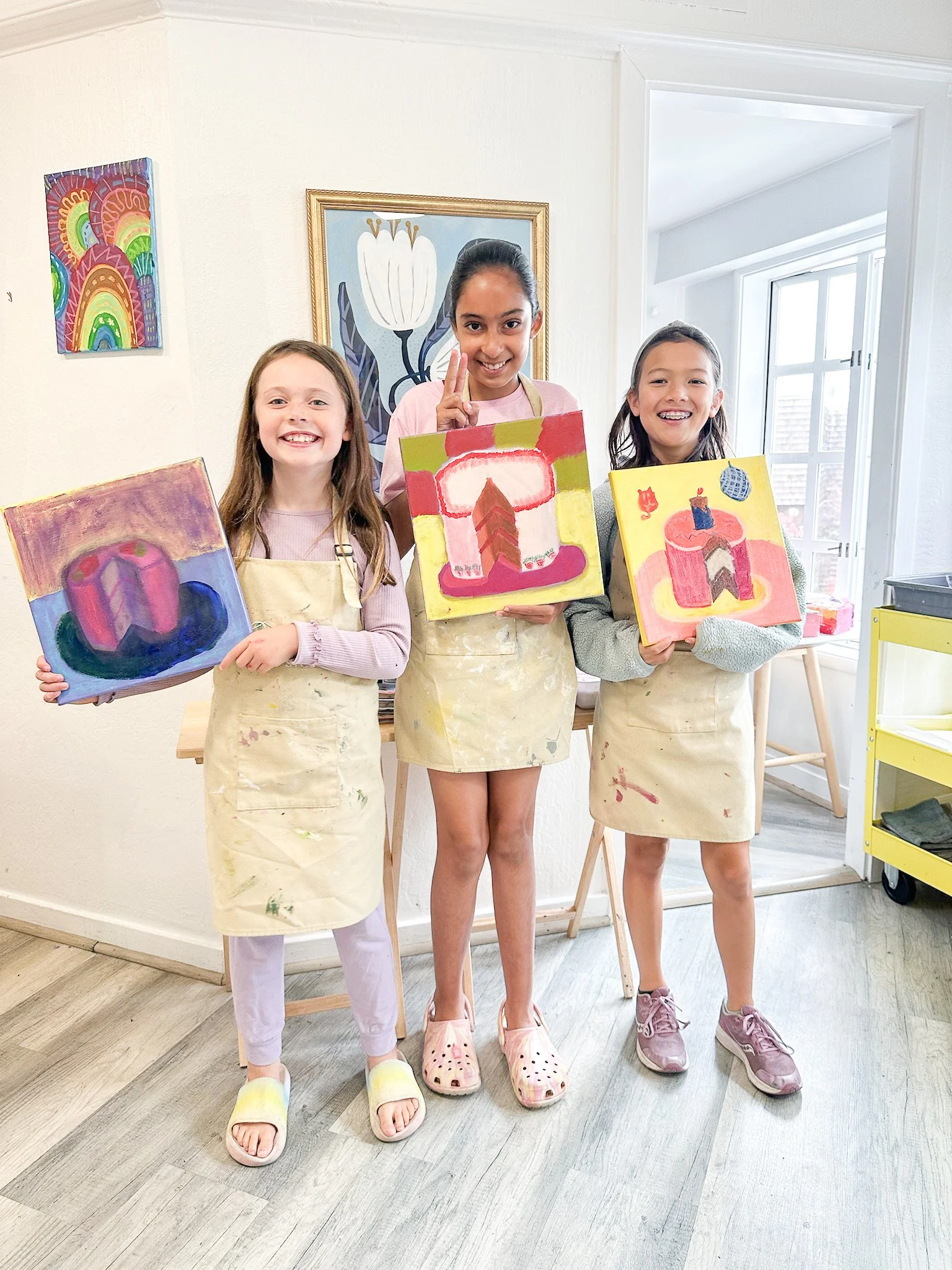 Three young girls in paint-stained aprons holding colorful abstract paintings in an art studio