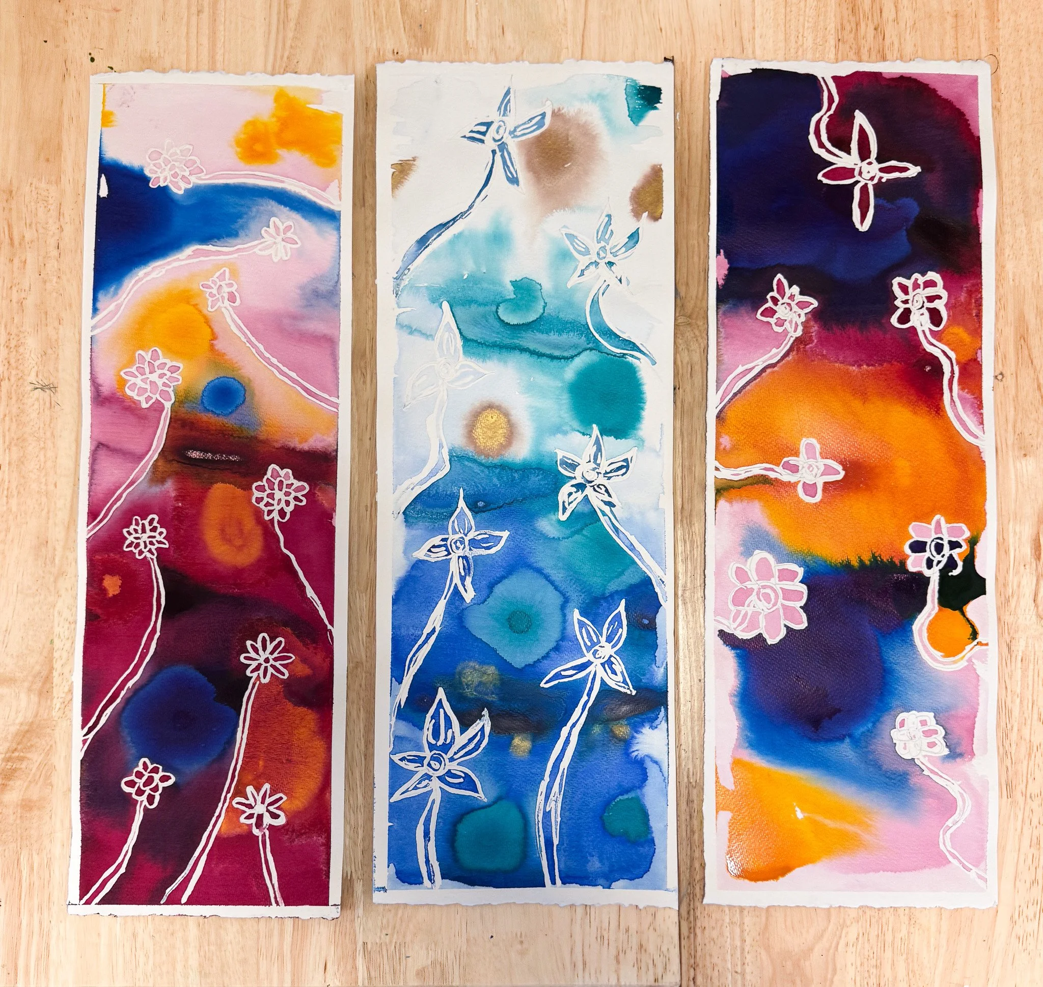 Three watercolor paintings on paper with abstract floral designs, each featuring white stenciled flowers over colorful backgrounds of pink, blue, orange, and purple, laid on a wooden surface.