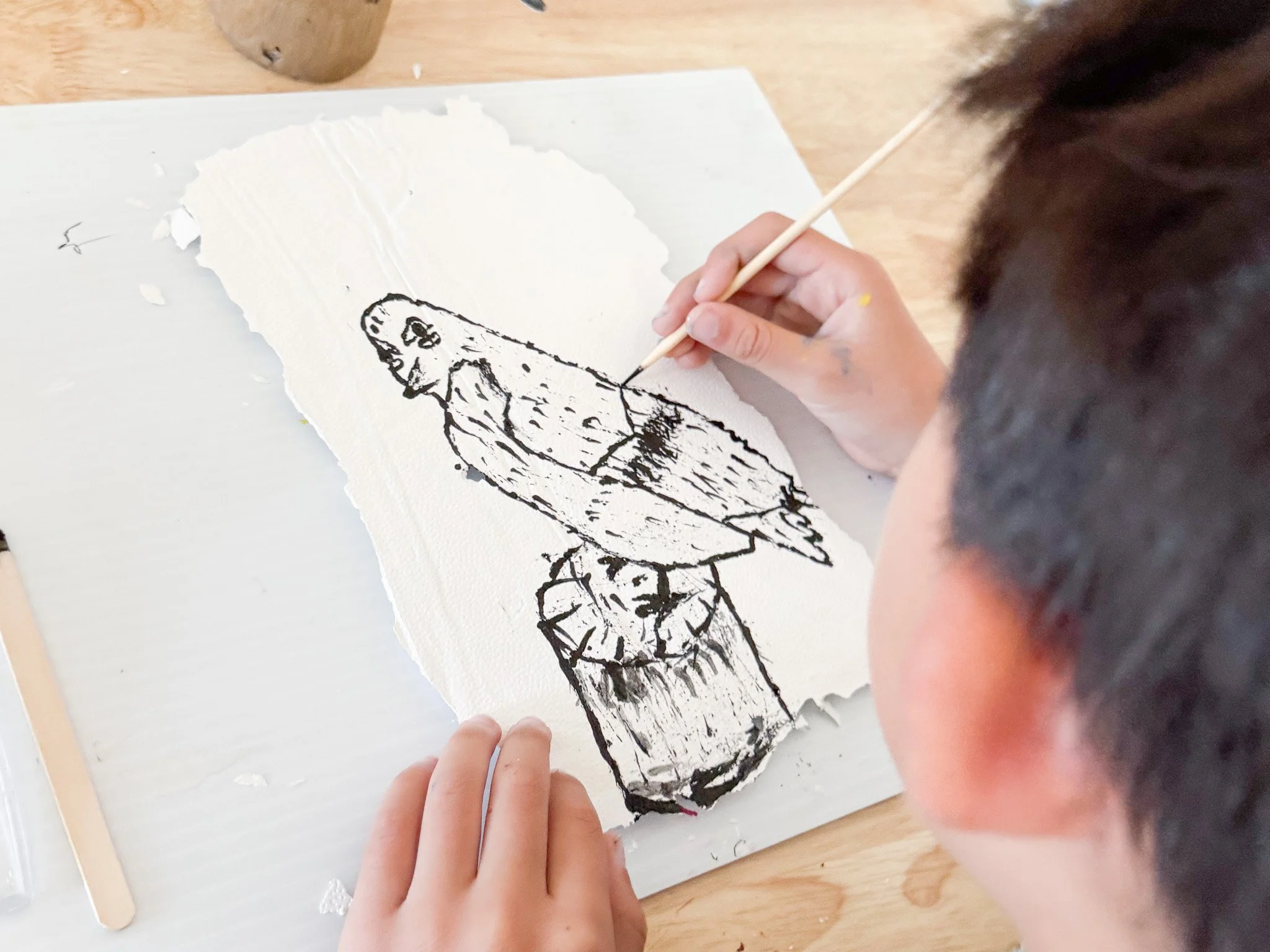 Child creating a black ink drawing of a bird on a torn piece of white paper.