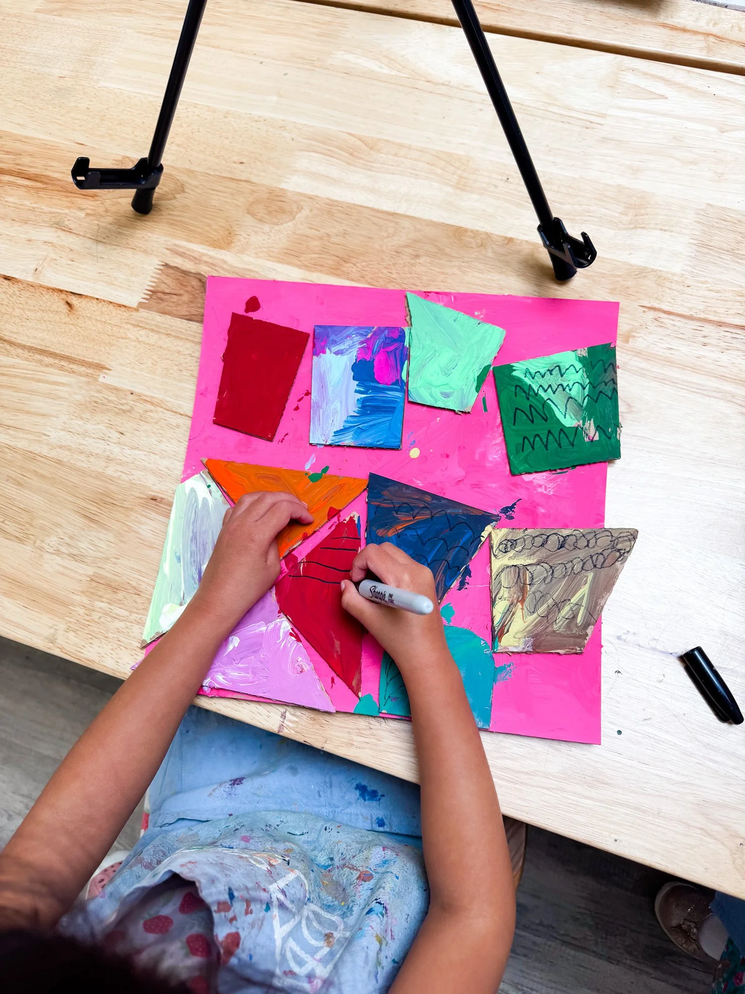 Child's hands drawing colorful painted paper shapes on pink poster board.