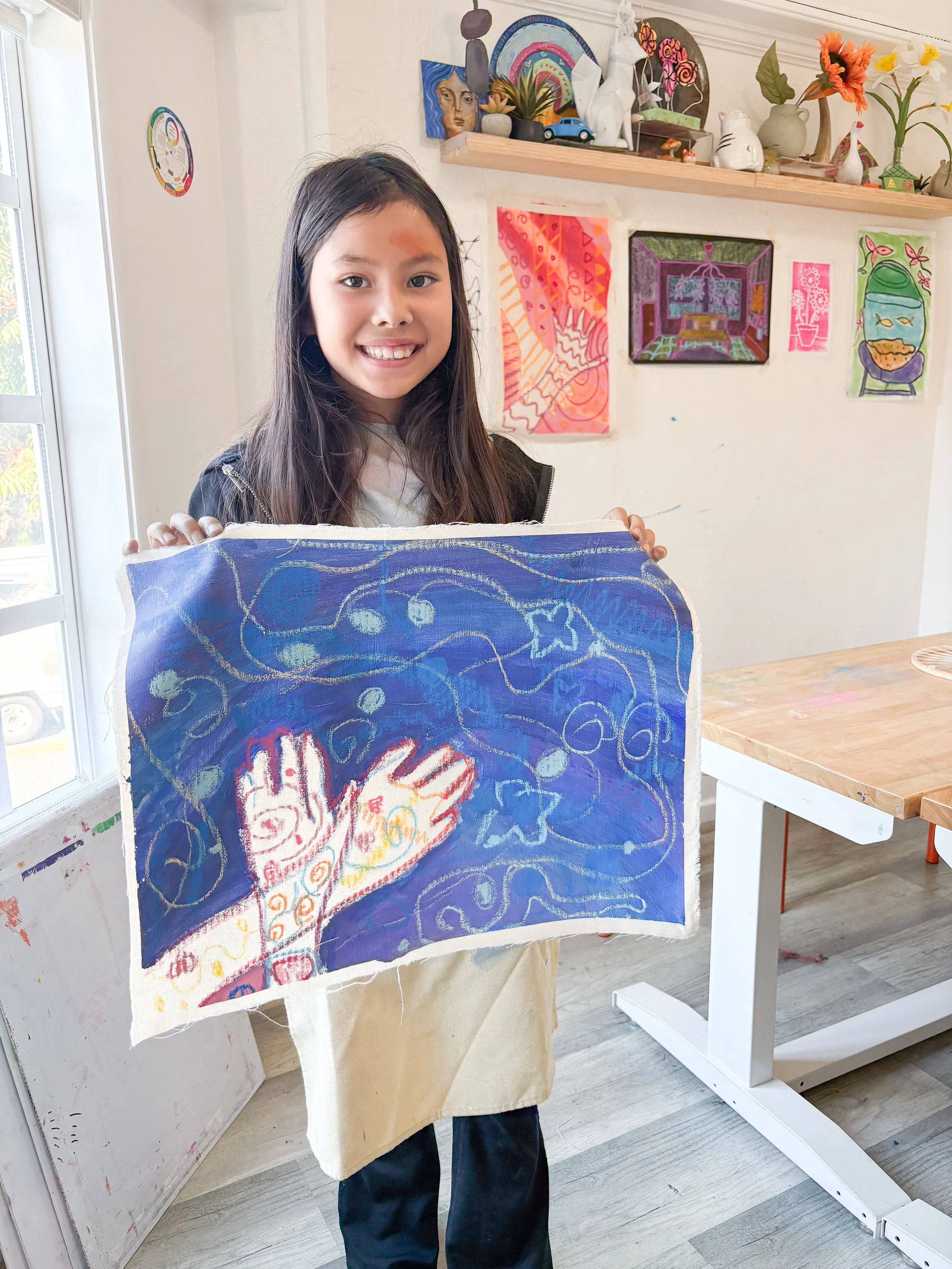 A young girl with long dark hair smiling and holding up a colorful drawing or painting while standing in an art studio or classroom. The artwork features a hand with abstract, vibrant patterns on a blue background.