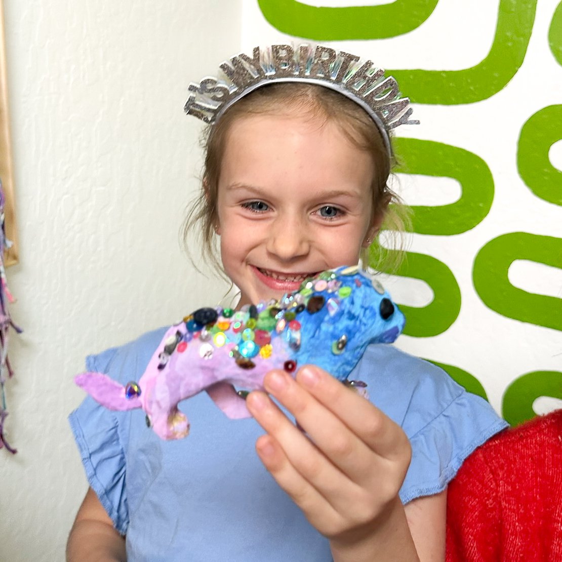 A young girl smiling and holding a colorful craft project, wearing a birthday headband with silver letters, in front of a white and green patterned wall.
