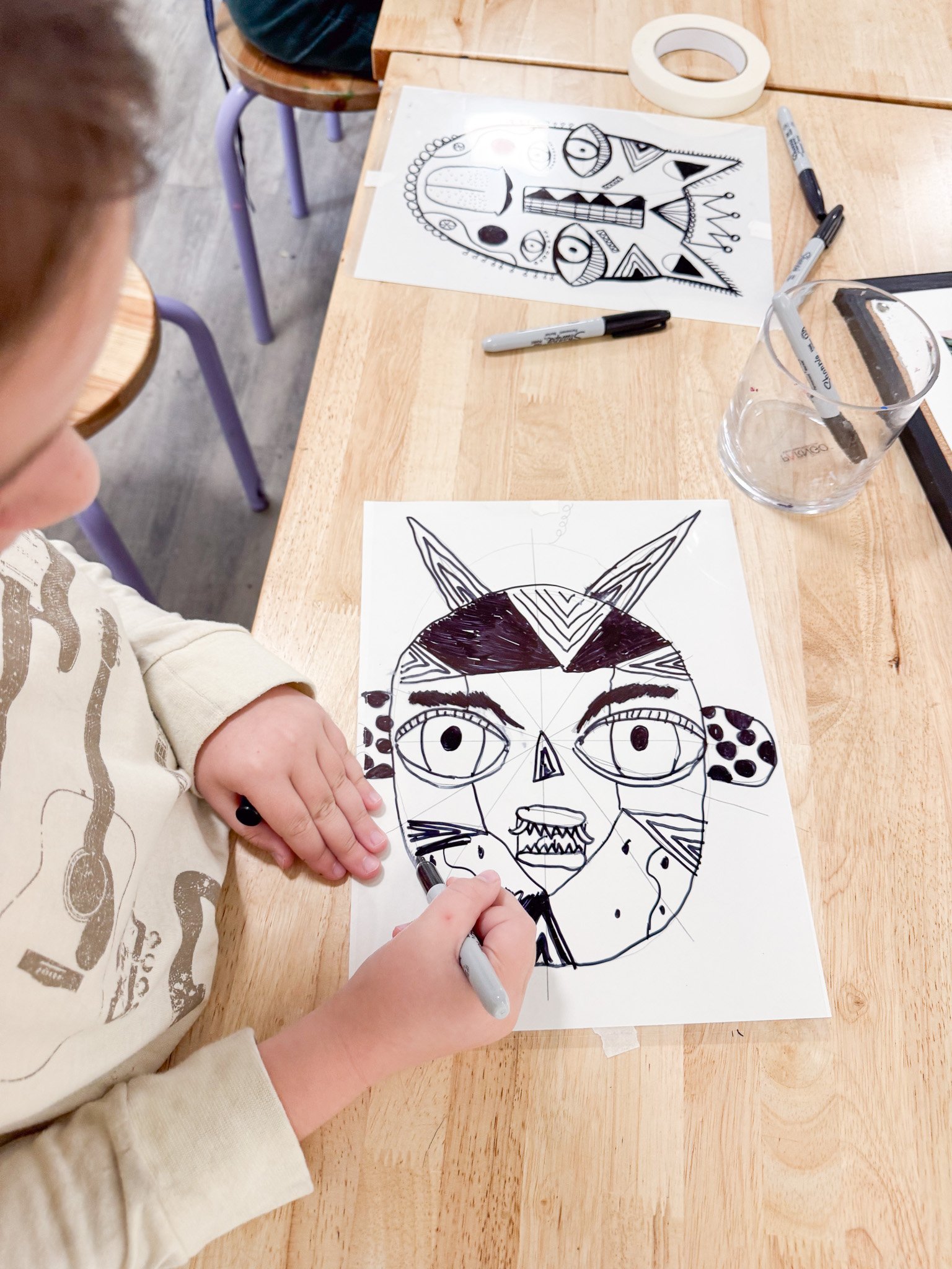 Child drawing a black and white abstract face with large eyes, ears, horns, and sharp features on paper at a wooden table, with art supplies and artwork in the background.