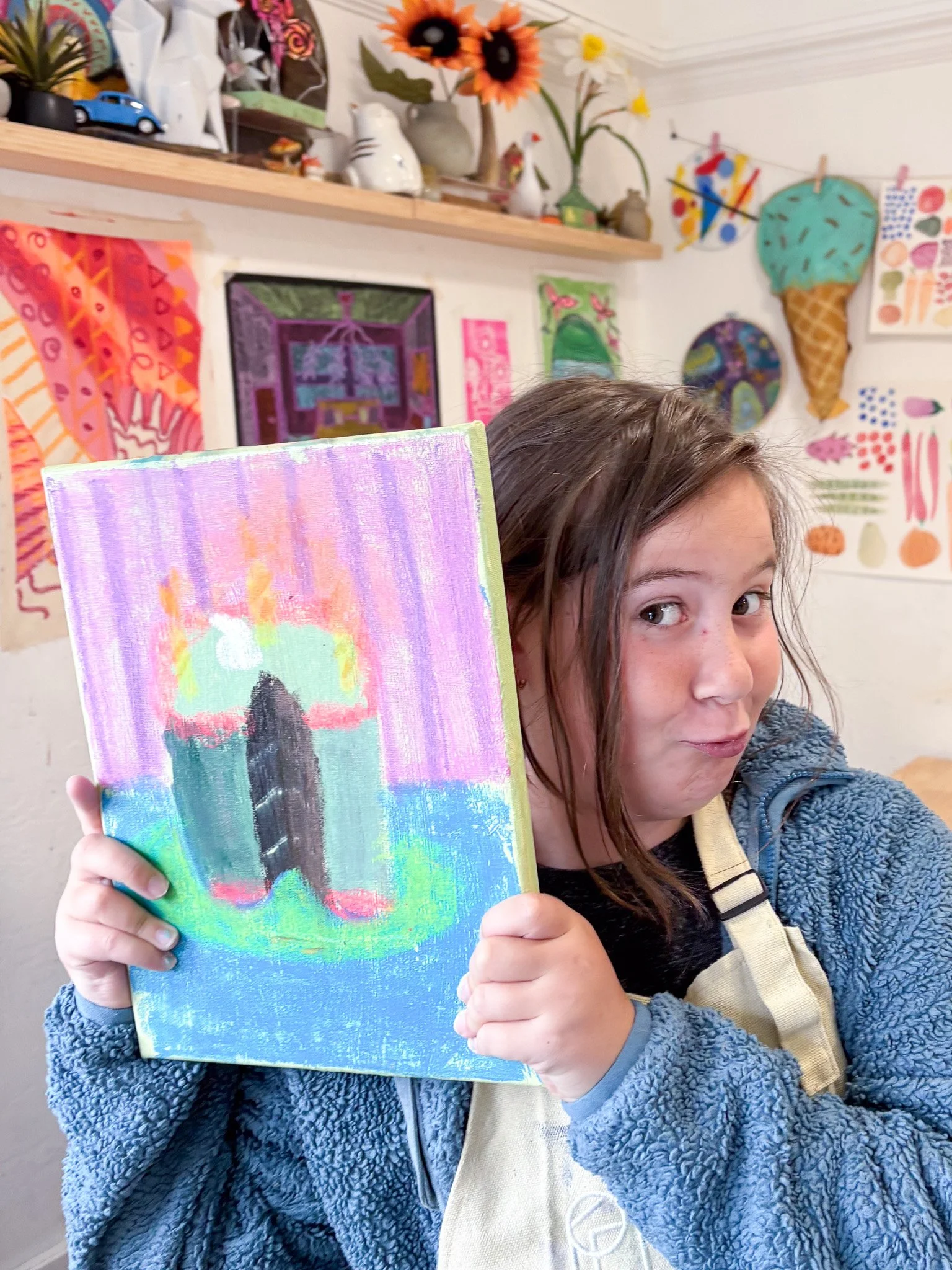 Young girl holding up a colorful painting of a rocket with a sunset background in an art studio with various artworks and decorations.