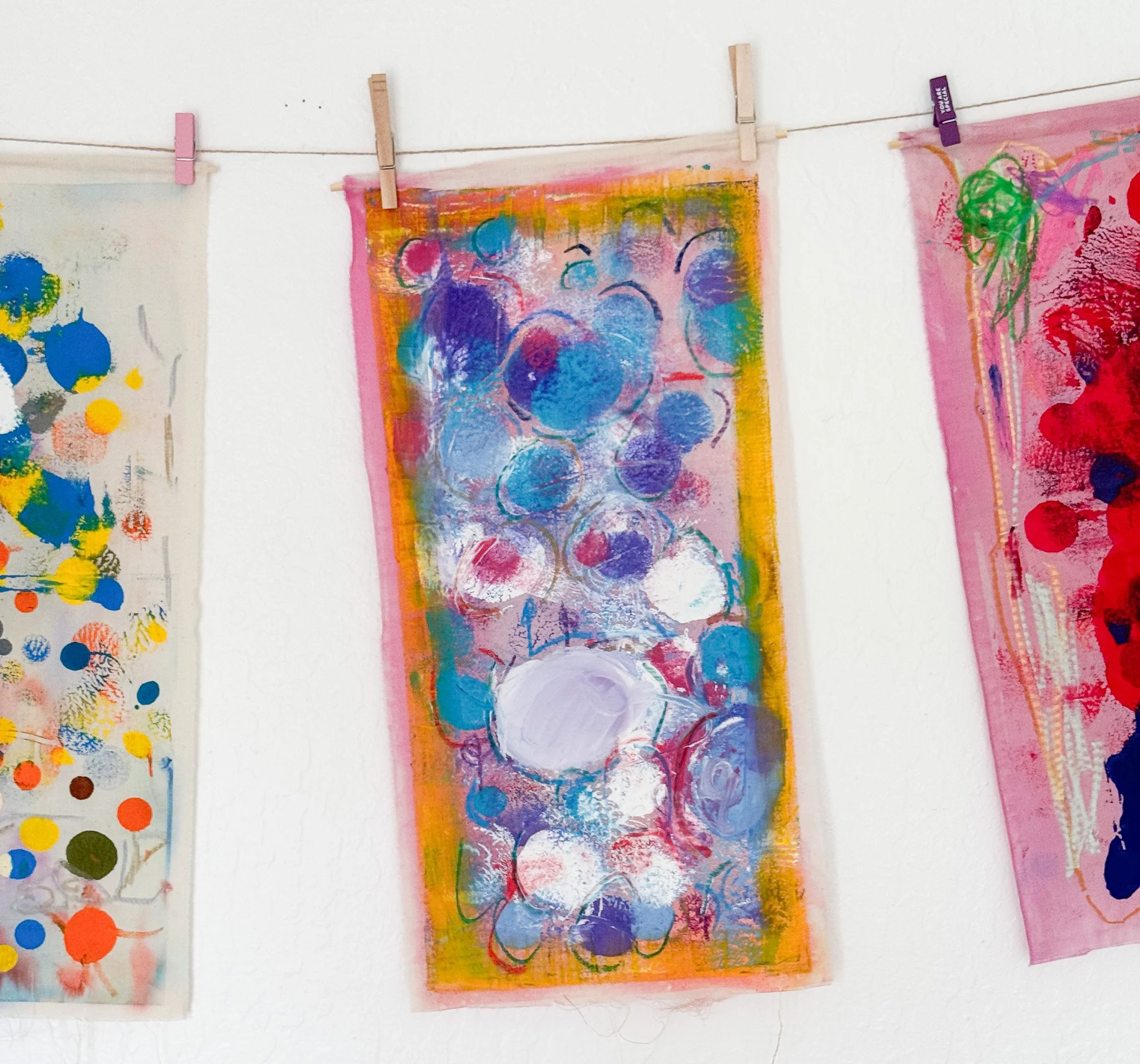 Colorful abstract art on fabric with circles, splashes, and strokes, hung on a wire with clothespins.