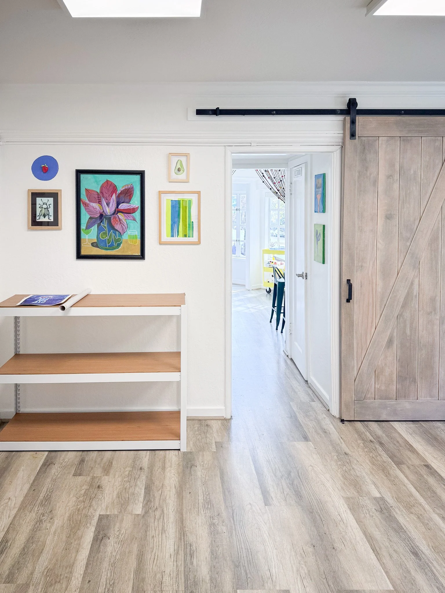 Interior view of a room with wood flooring, a white wall decorated with framed artwork, a white shelf, and a sliding barn door. The barn door is partially open, revealing another room with a dining table and chairs.