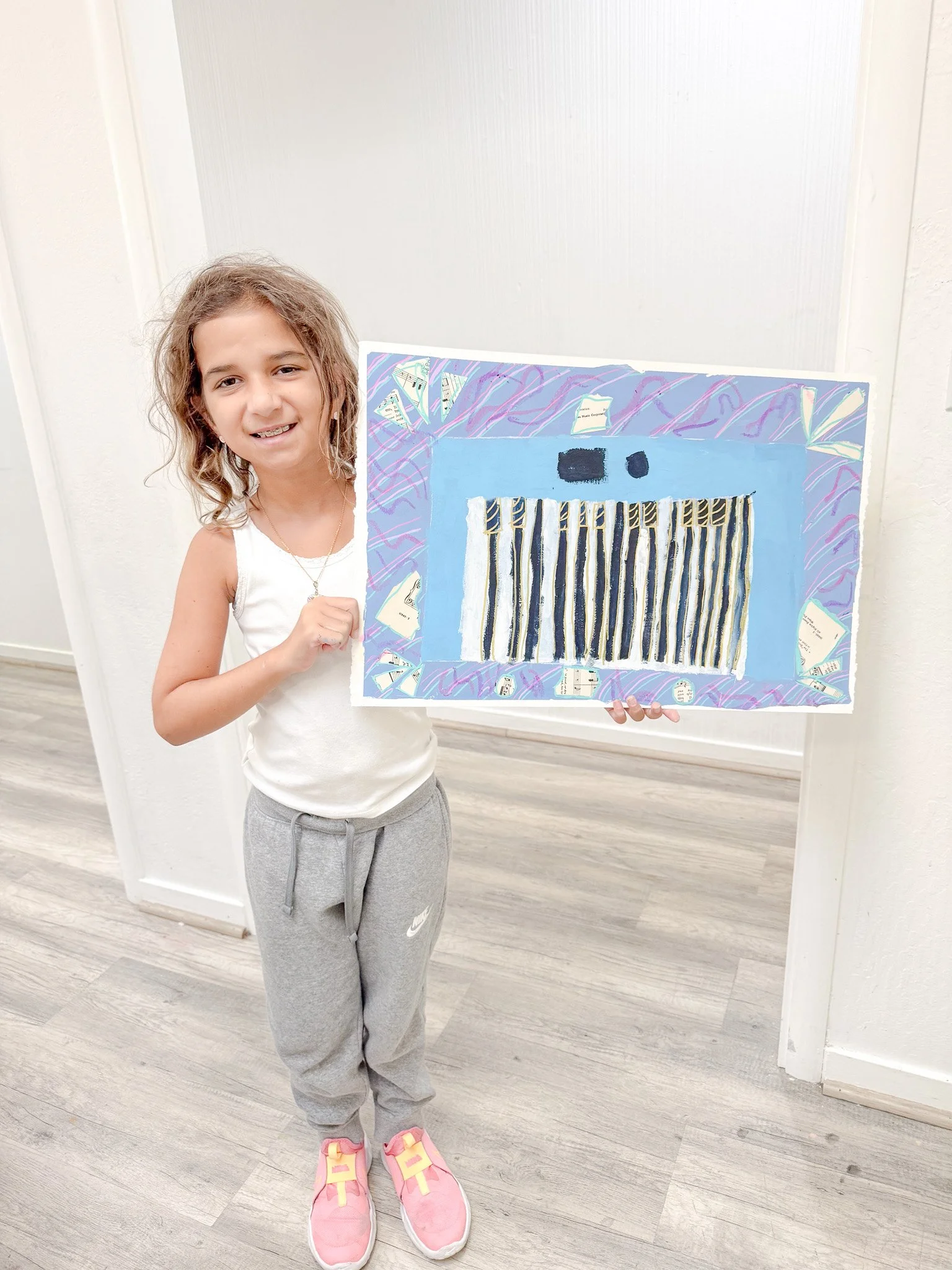 Young girl smiling and holding a colorful abstract artwork on paper, standing indoors on light wood flooring, wearing a white sleeveless top, gray sweatpants, and pink sneakers.