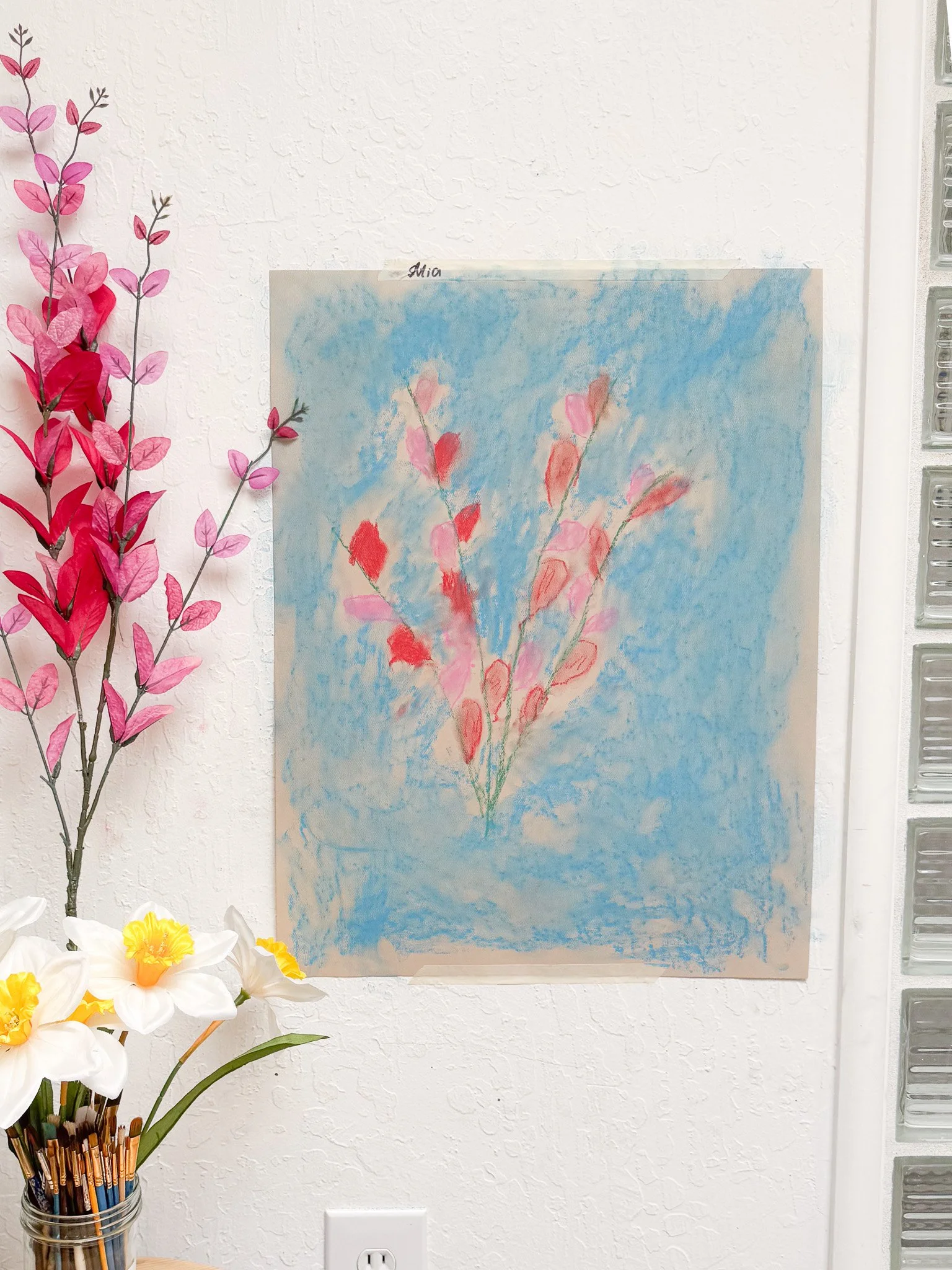 A child's drawing of pink flowers on a blue background, taped to a white wall.