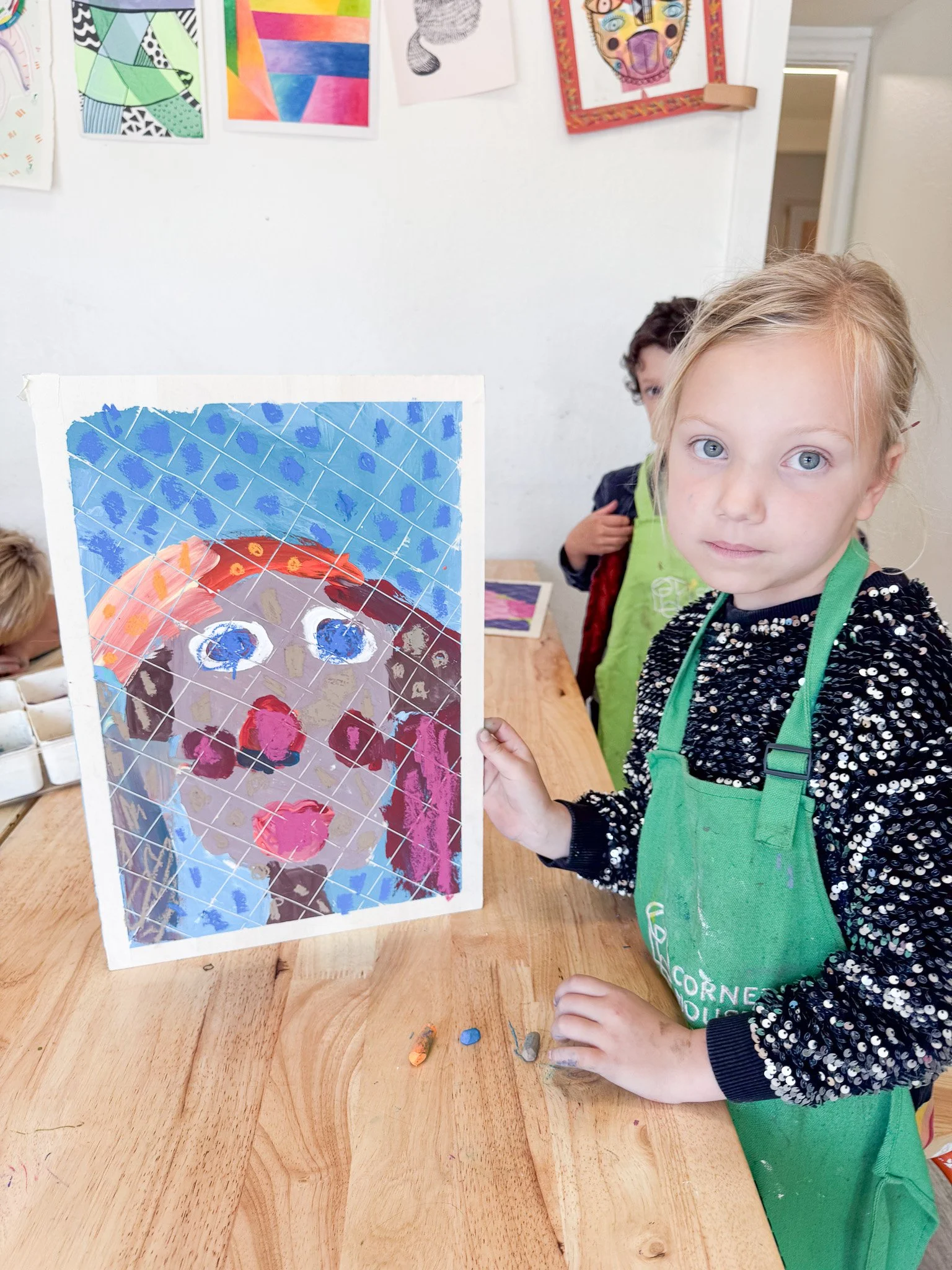 Young girl with blonde hair holding an abstract colorful artwork of a face painted on a clear sheet, with other children in the background, in an art classroom.