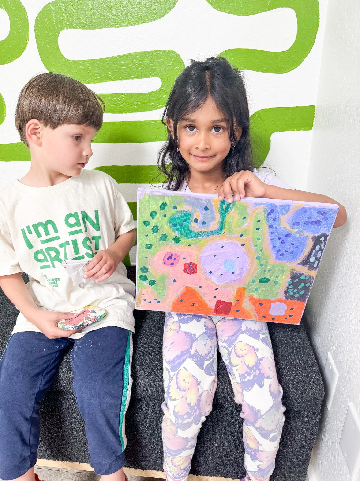 A young girl sitting on a gray bench, holding up a colorful abstract painting, with a boy sitting beside her looking at the painting. They are in an indoor setting with a white wall and green wall art behind them.