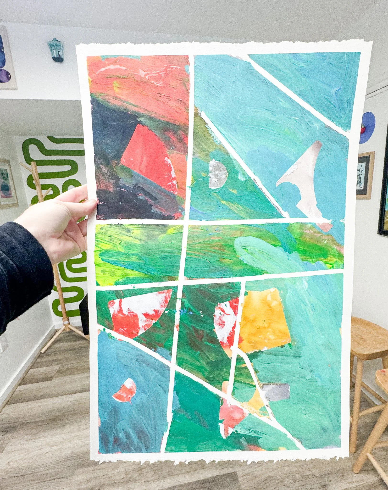 Hand holding a colorful abstract painting divided into nine sections with various brushstrokes and shapes.