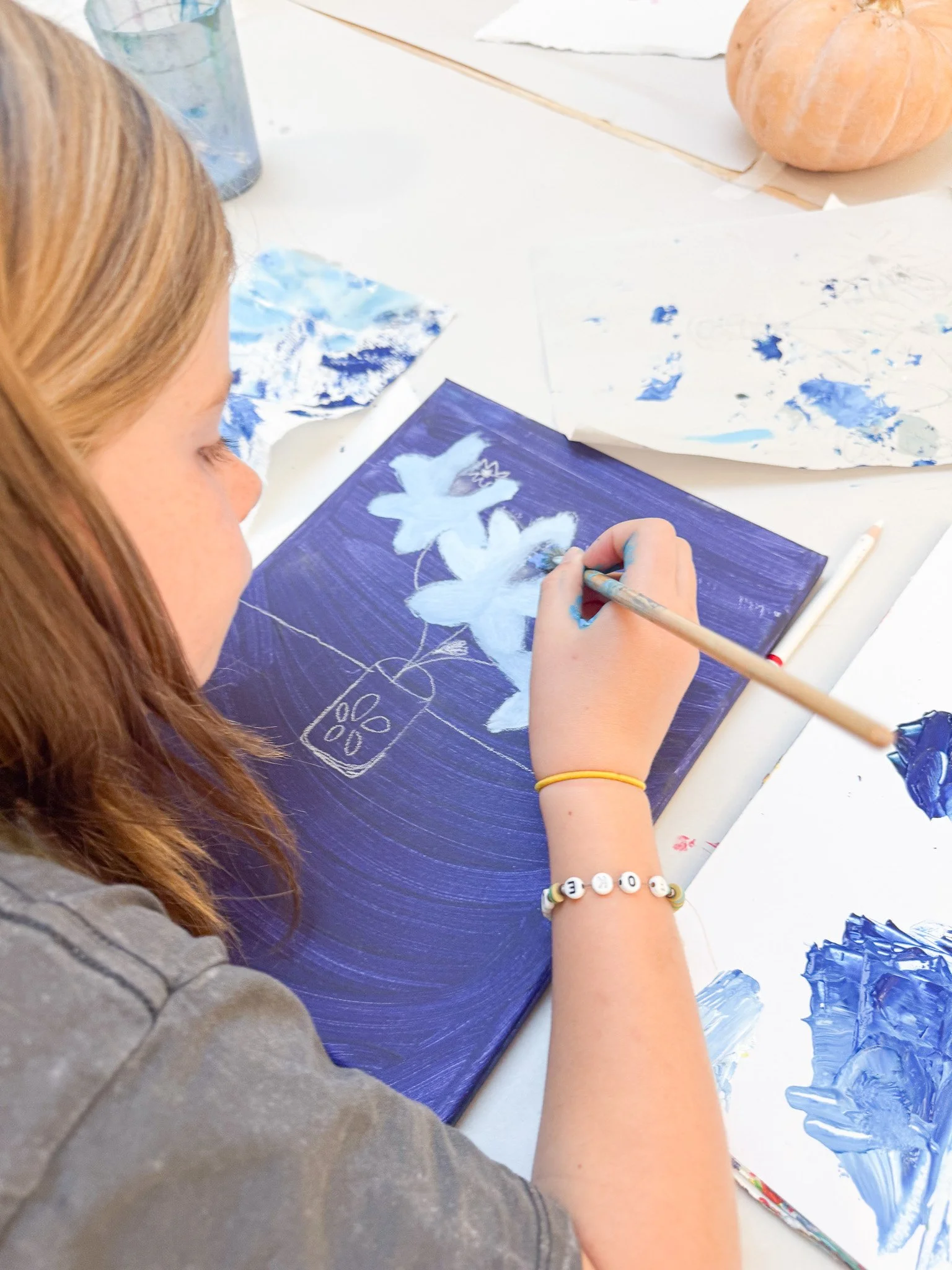 A girl with reddish-brown hair painting a floral design on a dark blue canvas using white paint, with various art supplies and a pumpkin on the table.