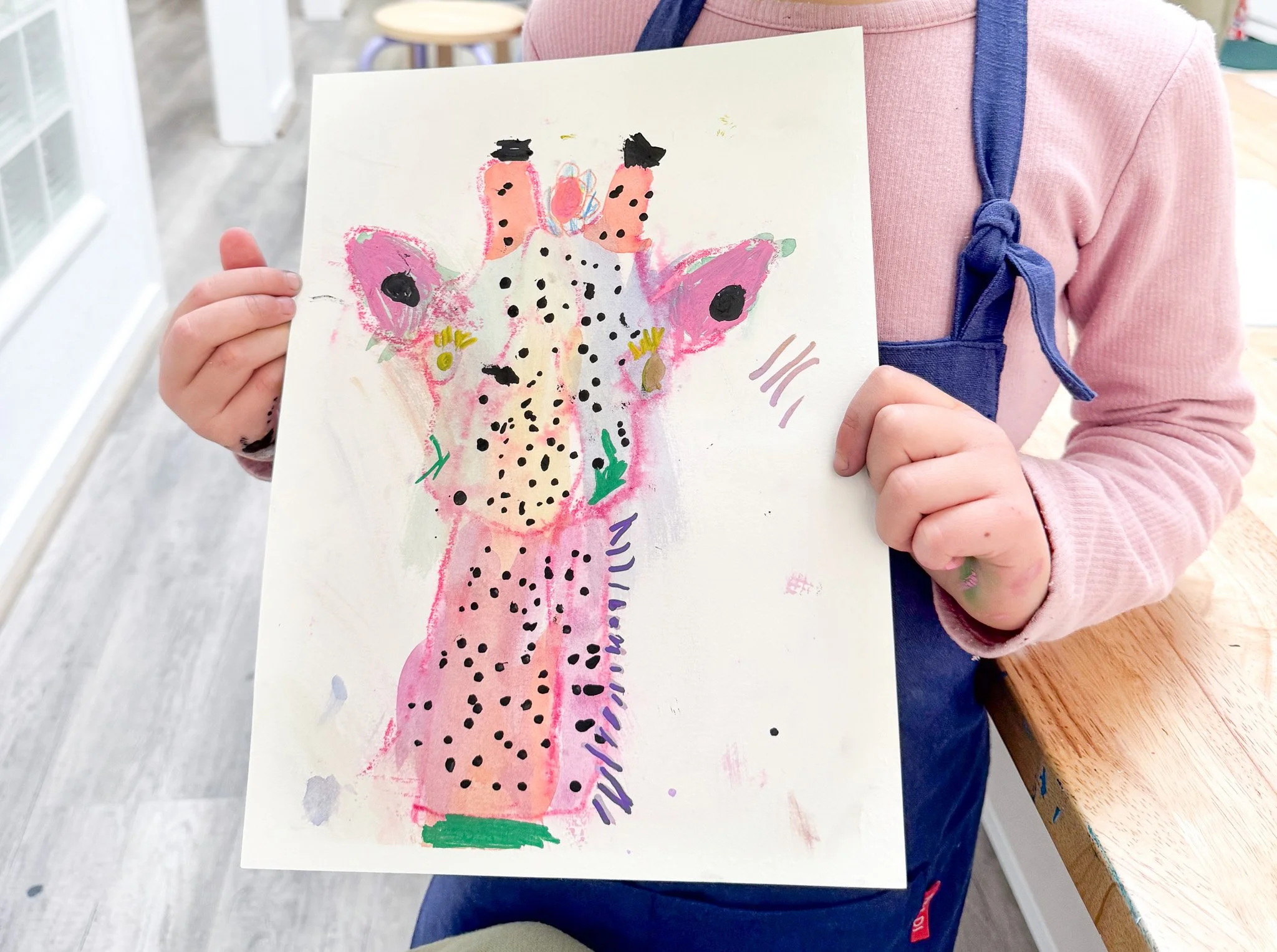 Child in pink sweater and blue apron holding a colorful, abstract watercolor painting of a giraffe.
