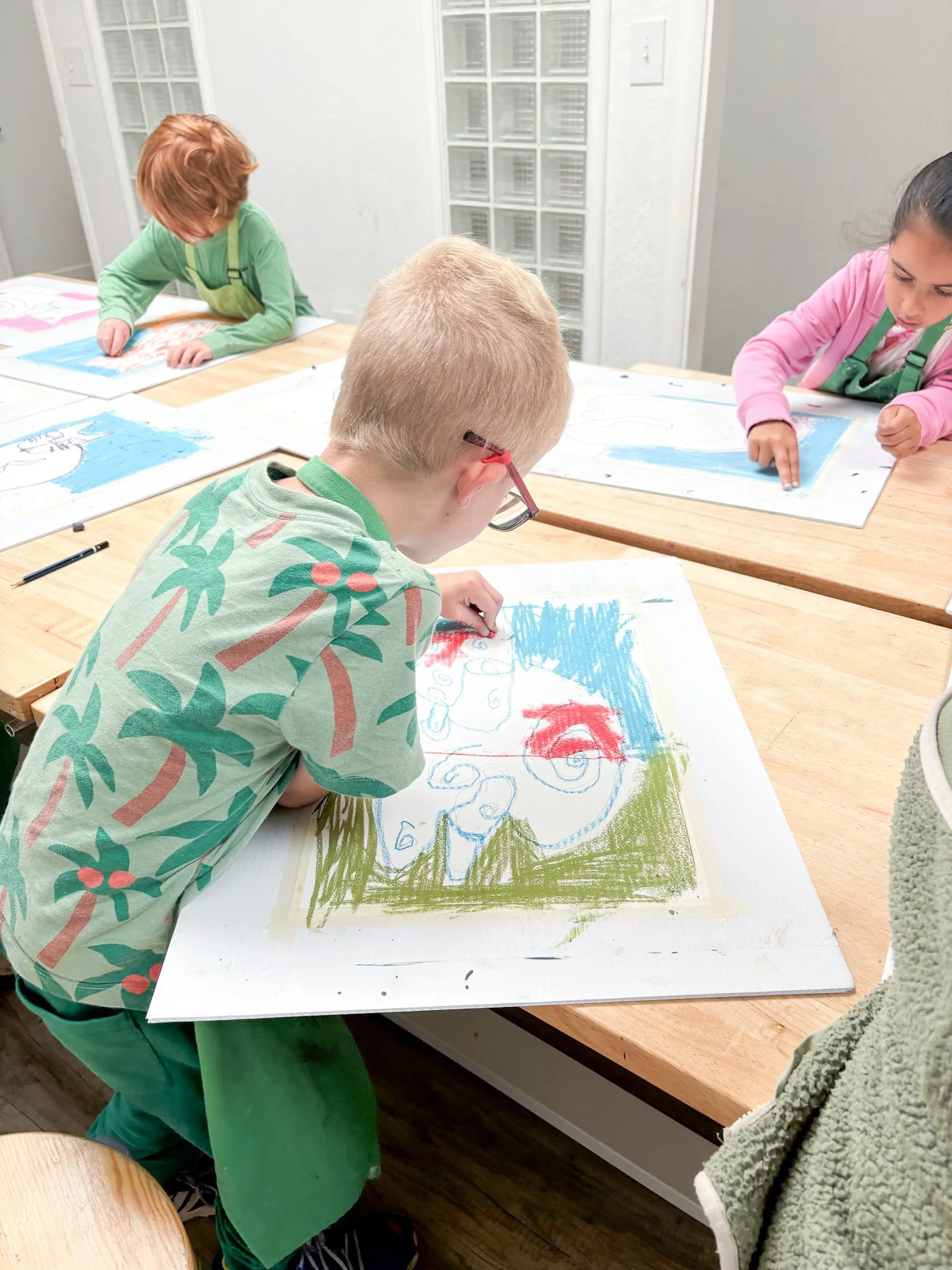 Children working on colorful abstract drawings at a wooden table in a classroom.