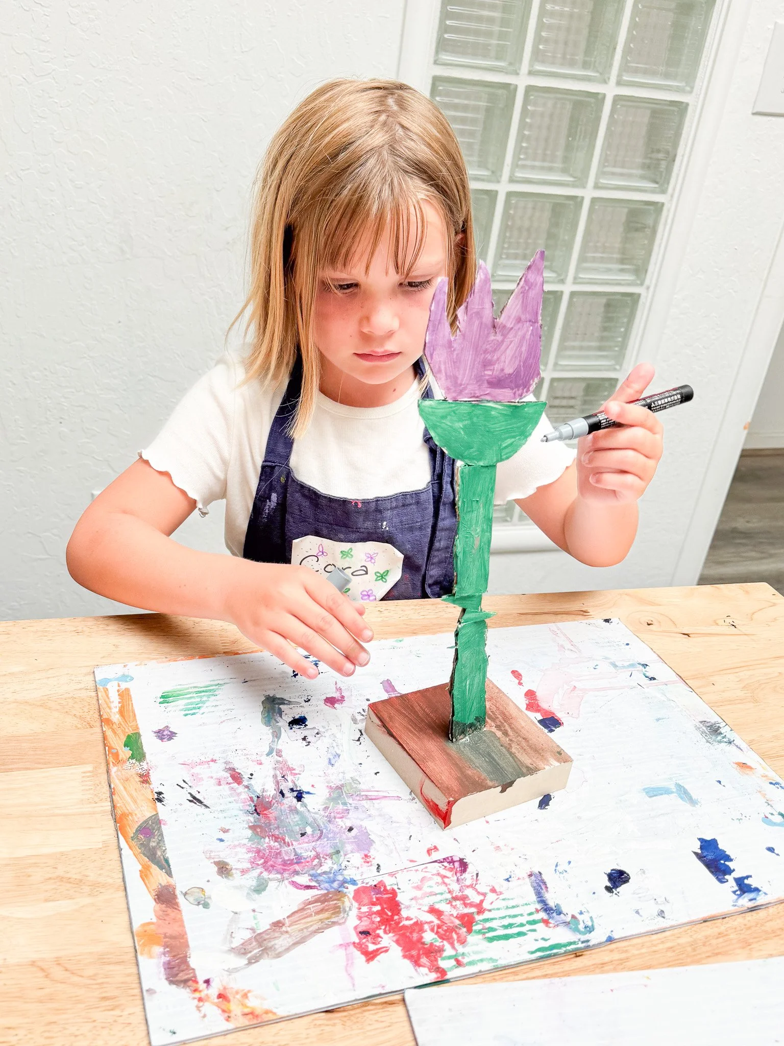A young girl with blonde hair is painting a colorful paper flower statue at a wooden table covered with a paint-spattered sheet. She is focused, holding a black marker, and wearing a white shirt and a dark apron.