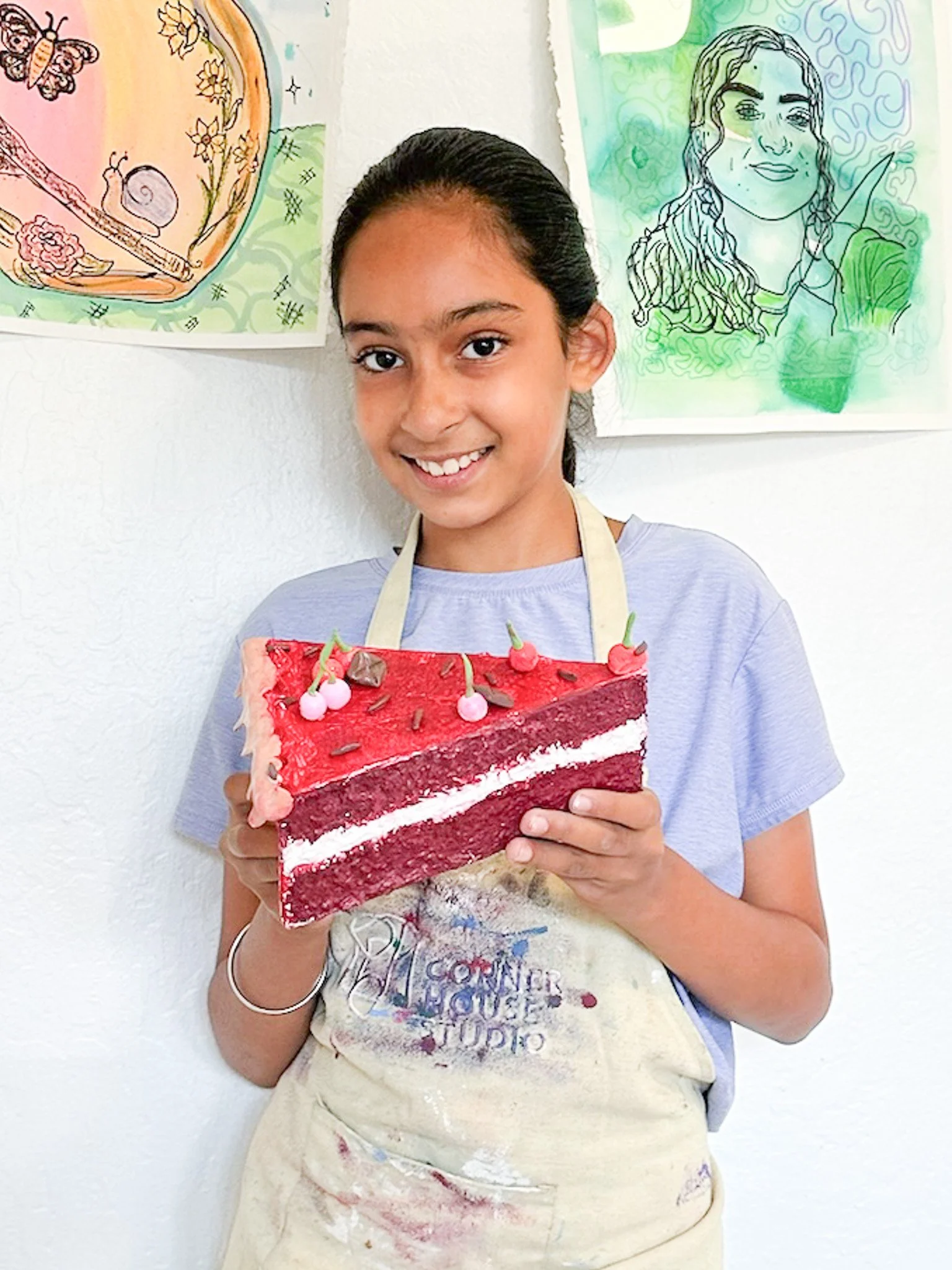 A young girl holding a slice of red velvet cake decorated with pink and red frosting and small decorative candies, smiling at the camera, standing in front of colorful artwork on a white wall.