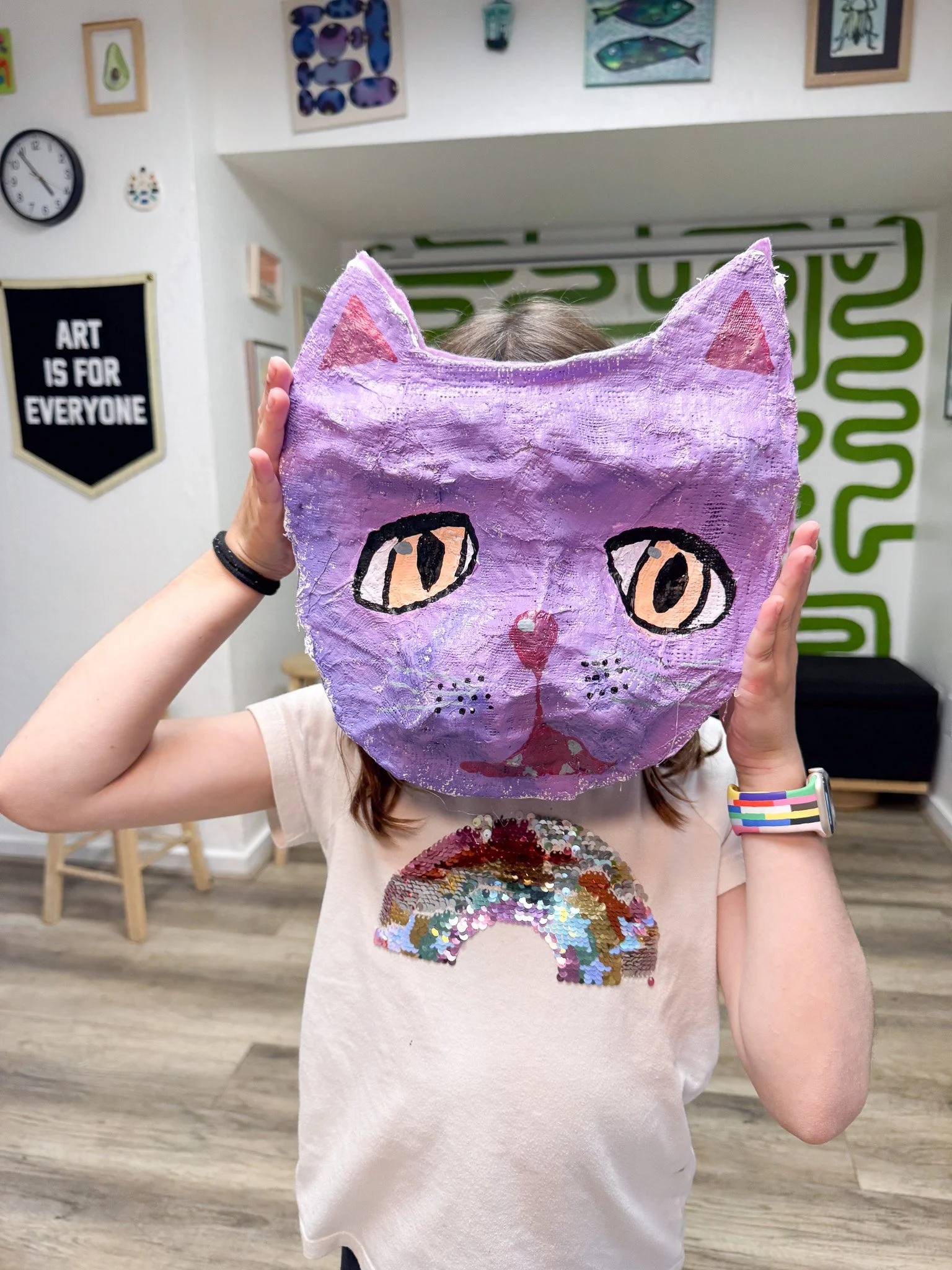 Child holding a purple cat mask in front of their face, with a rainbow sequin shirt, in a colorful art room.