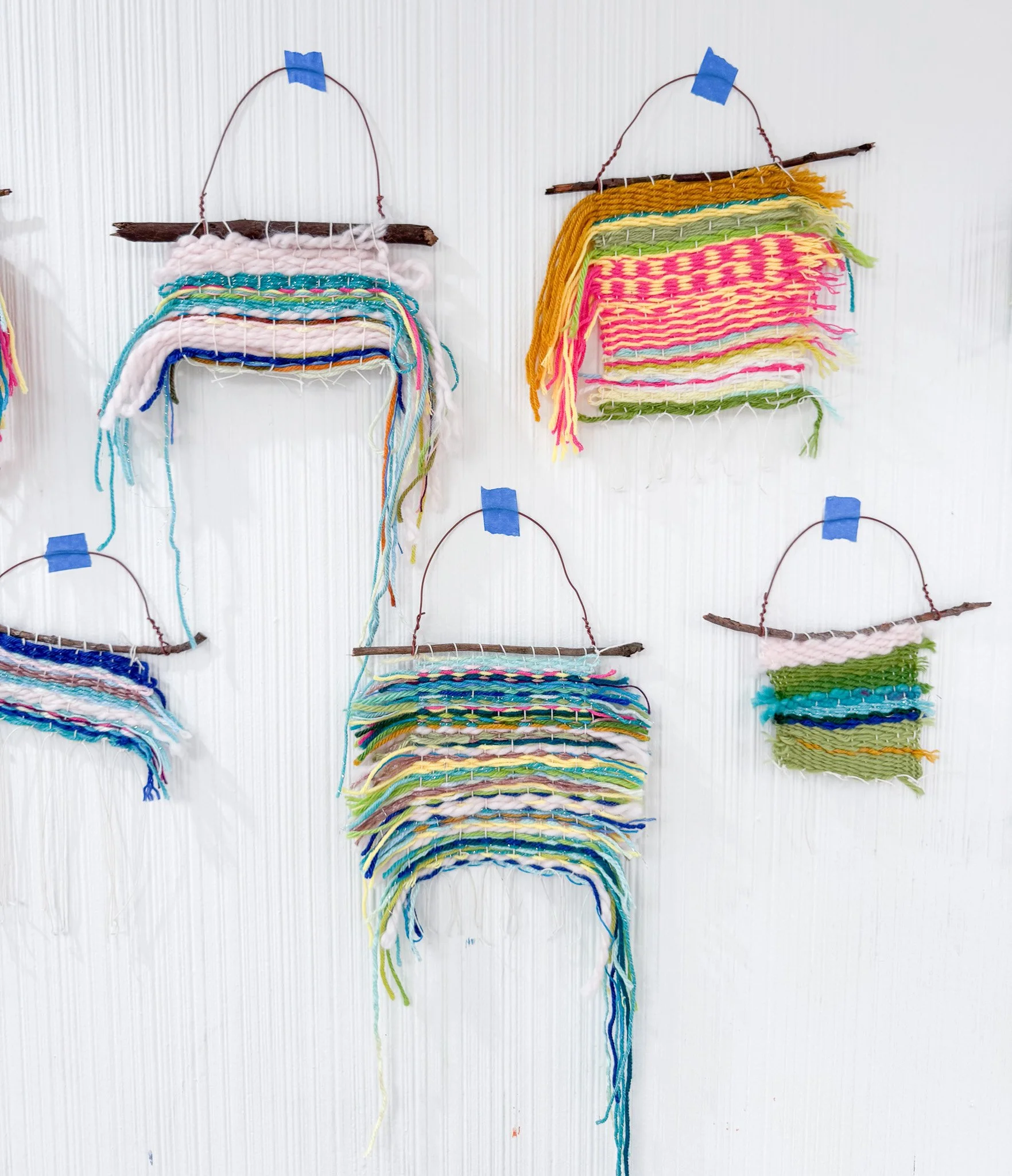Colorful woven wall hangings made with yarn and sticks, hung on a white wall with blue tape.