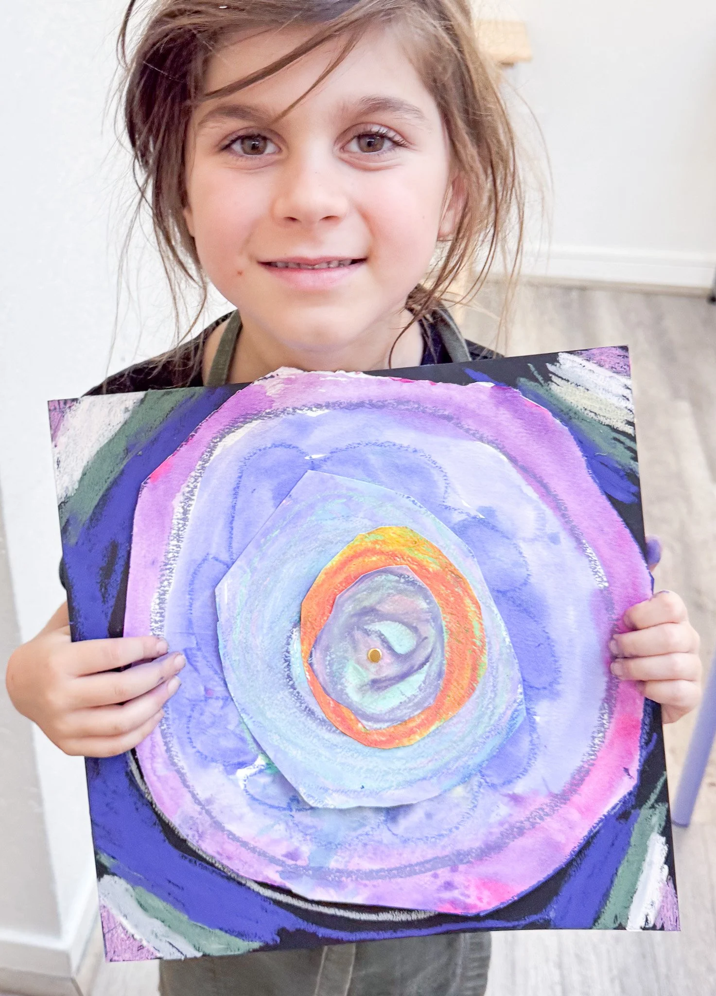 A young girl with brown hair and a black shirt holding a colorful abstract painting of concentric circles in purple, blue, orange, and yellow.
