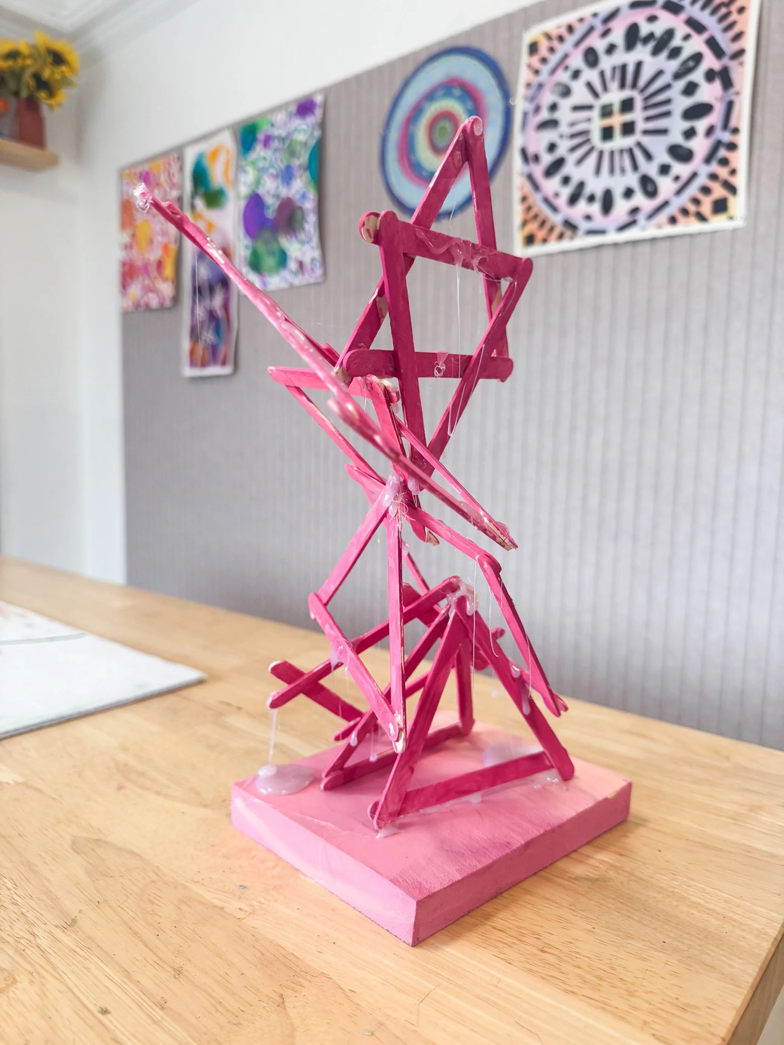 Pink star-shaped sculpture made of wooden sticks painted pink, mounted on a pink base, with colorful abstract artwork on the wall behind it.