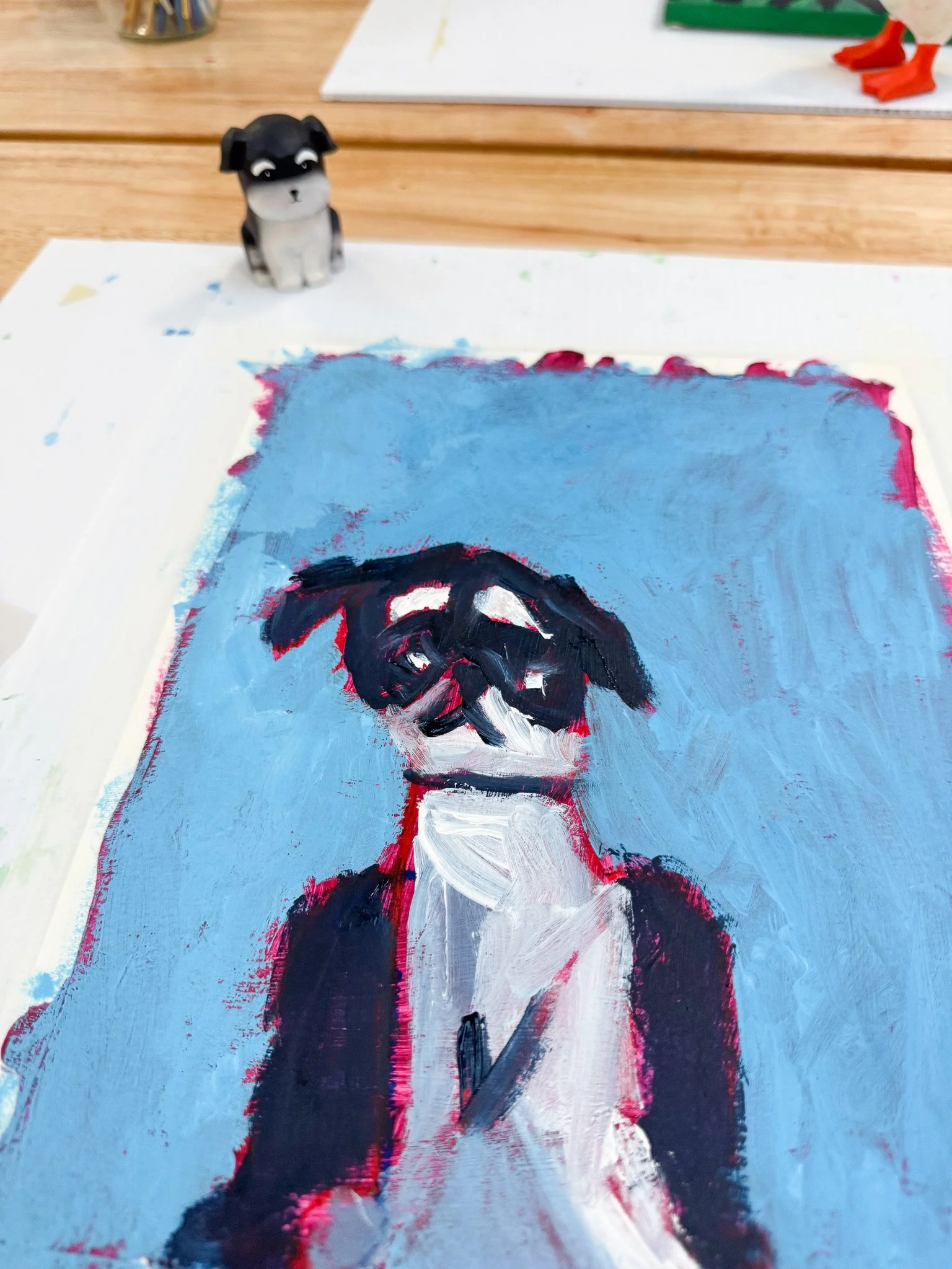 A painting of a black and white dog with a white face, wearing glasses, painted on a blue background. There is a small dog figurine on the table above the painting.