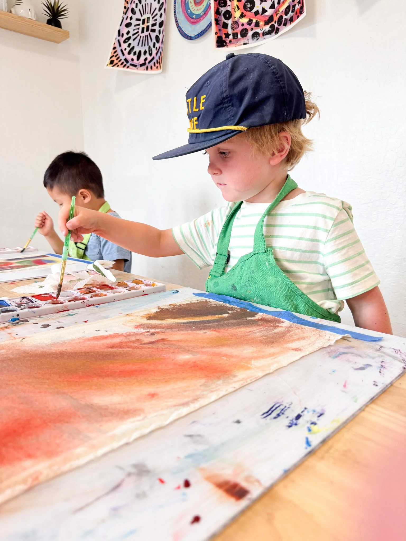 Two young boys painting with watercolors at a table, one wearing a blue cap and a green apron, with colorful artwork on the wall behind them.