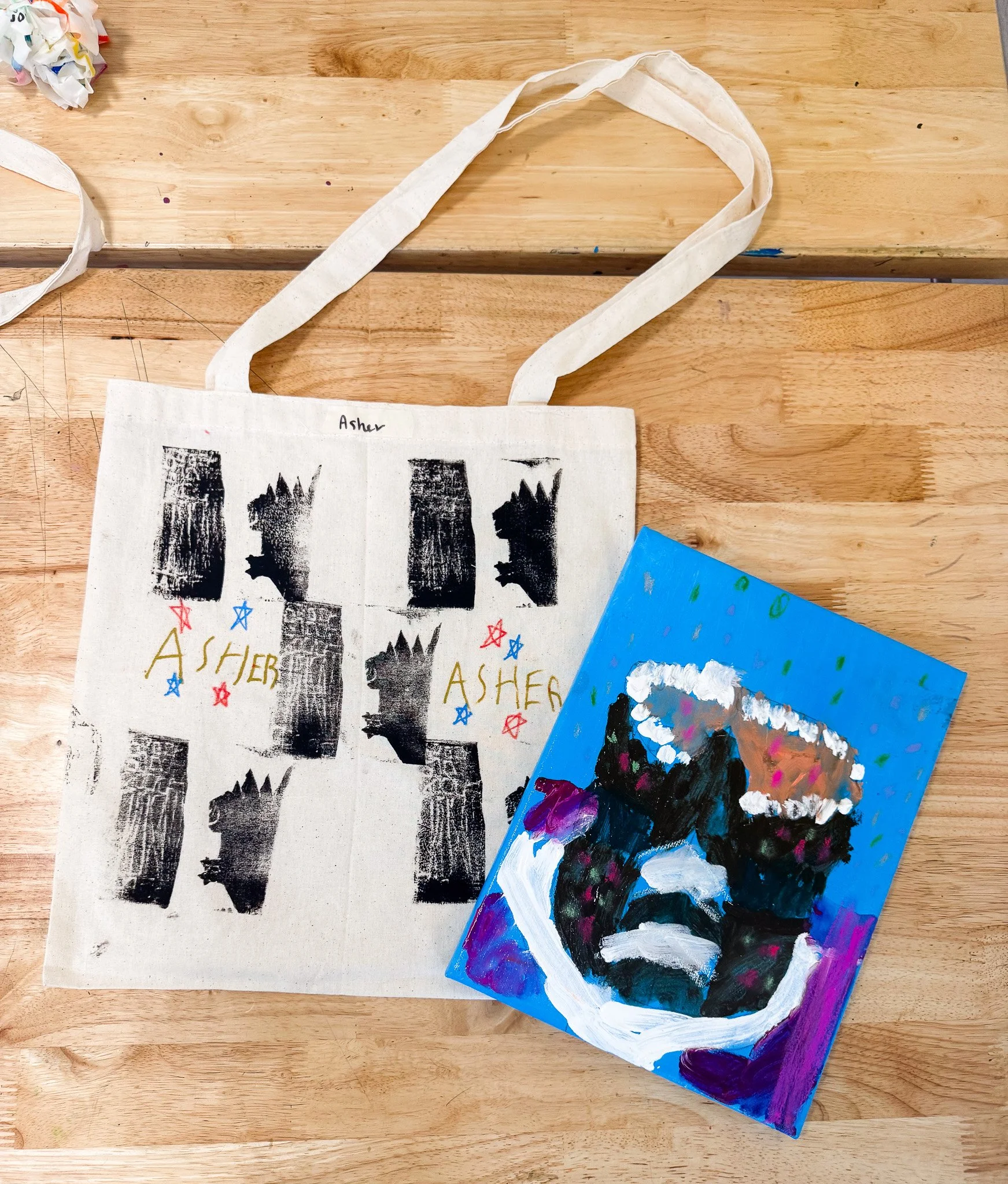 A tote bag with black ink prints of a dinosaur, decorated with colorful stars and the name 'Asher' written multiple times. Next to the bag, there is a colorful, abstract painting on a blue canvas featuring a large, dark face with vivid colors and a c