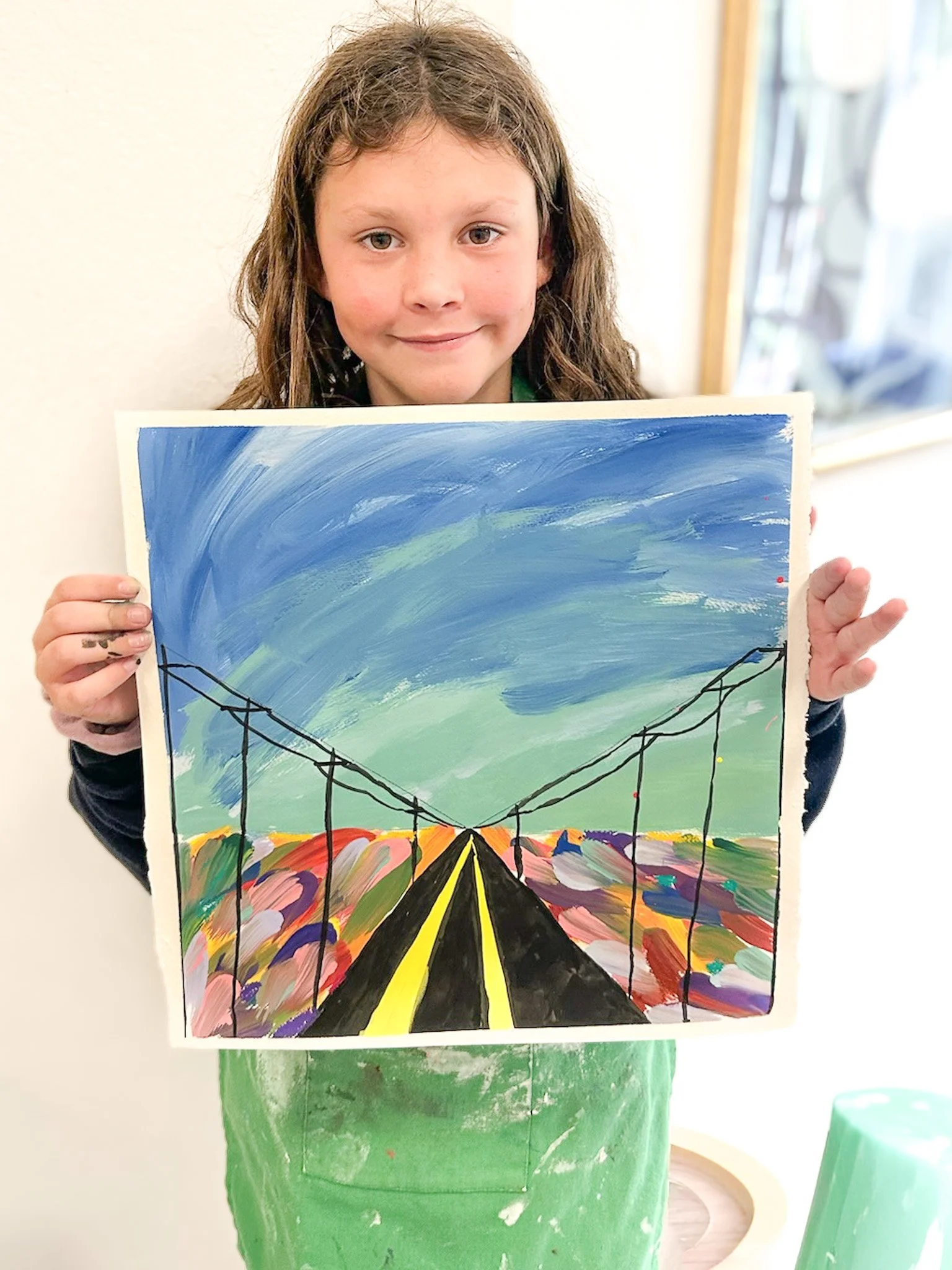 Young girl holding a colorful painting of a bridge with a sky background.