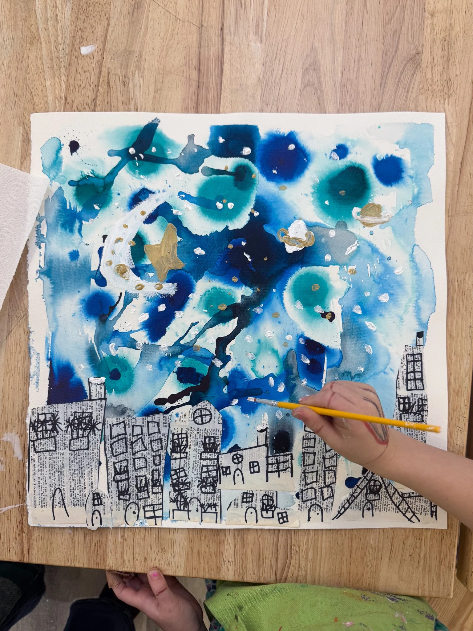 A child's hand painting a colorful abstract scene with black marker on a paper with cityscape outlines at the bottom, over a wooden table.