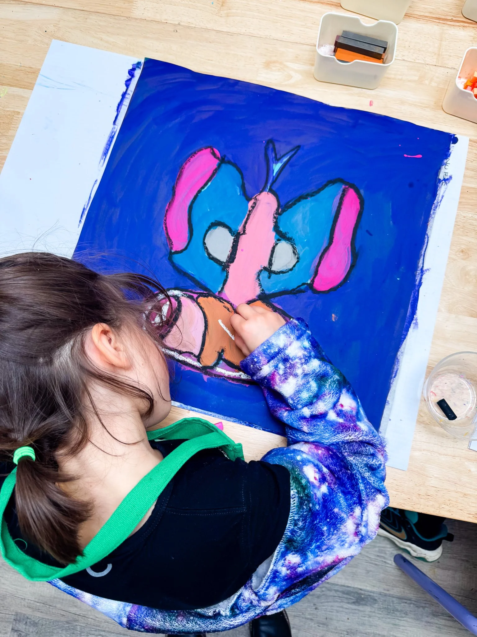 Child drawing and coloring a butterfly on a large blue sheet of paper, sitting at a wooden table with art supplies around.