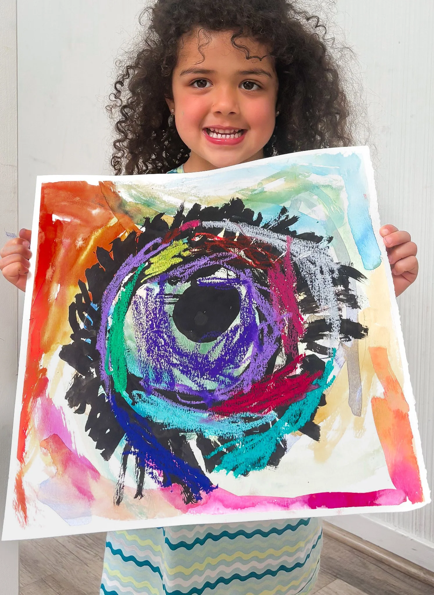Young girl with curly hair holding a colorful abstract painting of an eye, smiling.