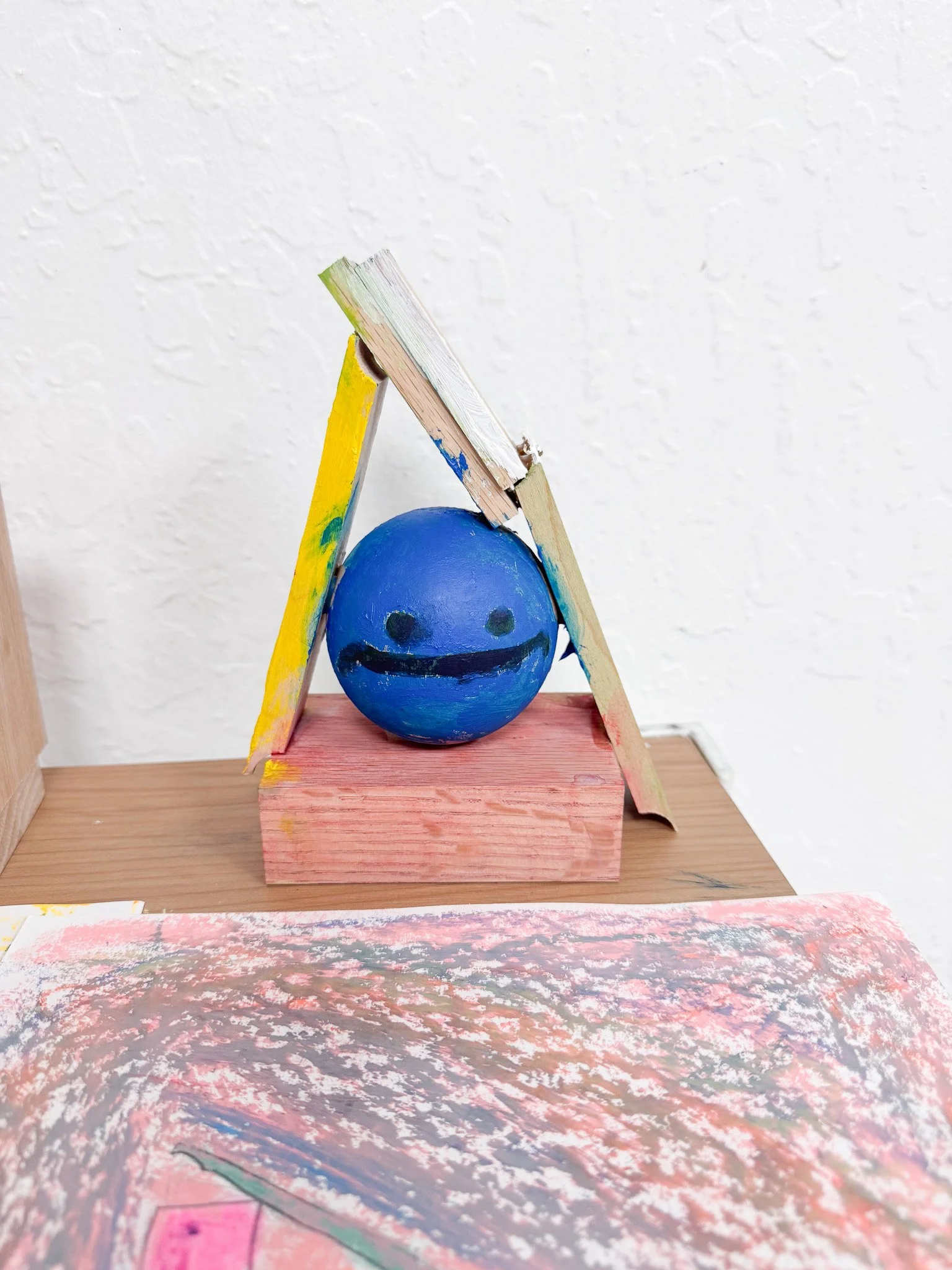 Blue smiling sphere with a black mouth and eyes, placed underneath a small wooden structure with painted multicolored planks, partly resting on a pink wooden block, set on a wooden surface.
