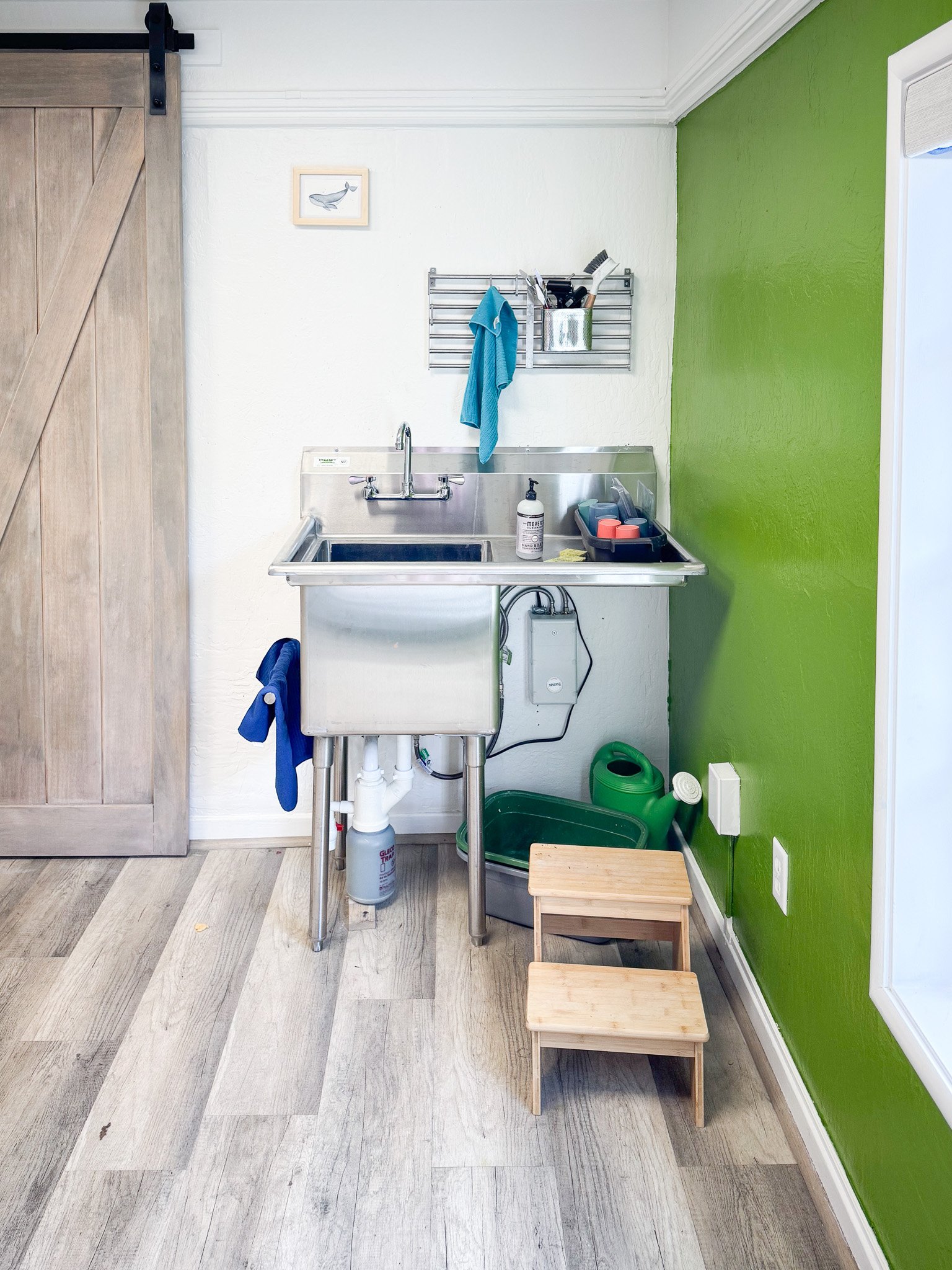 A small kitchen sink area with a stainless steel sink, green wall on the right, white wall behind, a wooden sliding door on the left, and a window with white trim. There are cleaning supplies, brushes, and an electric box under the sink, a turquoise 