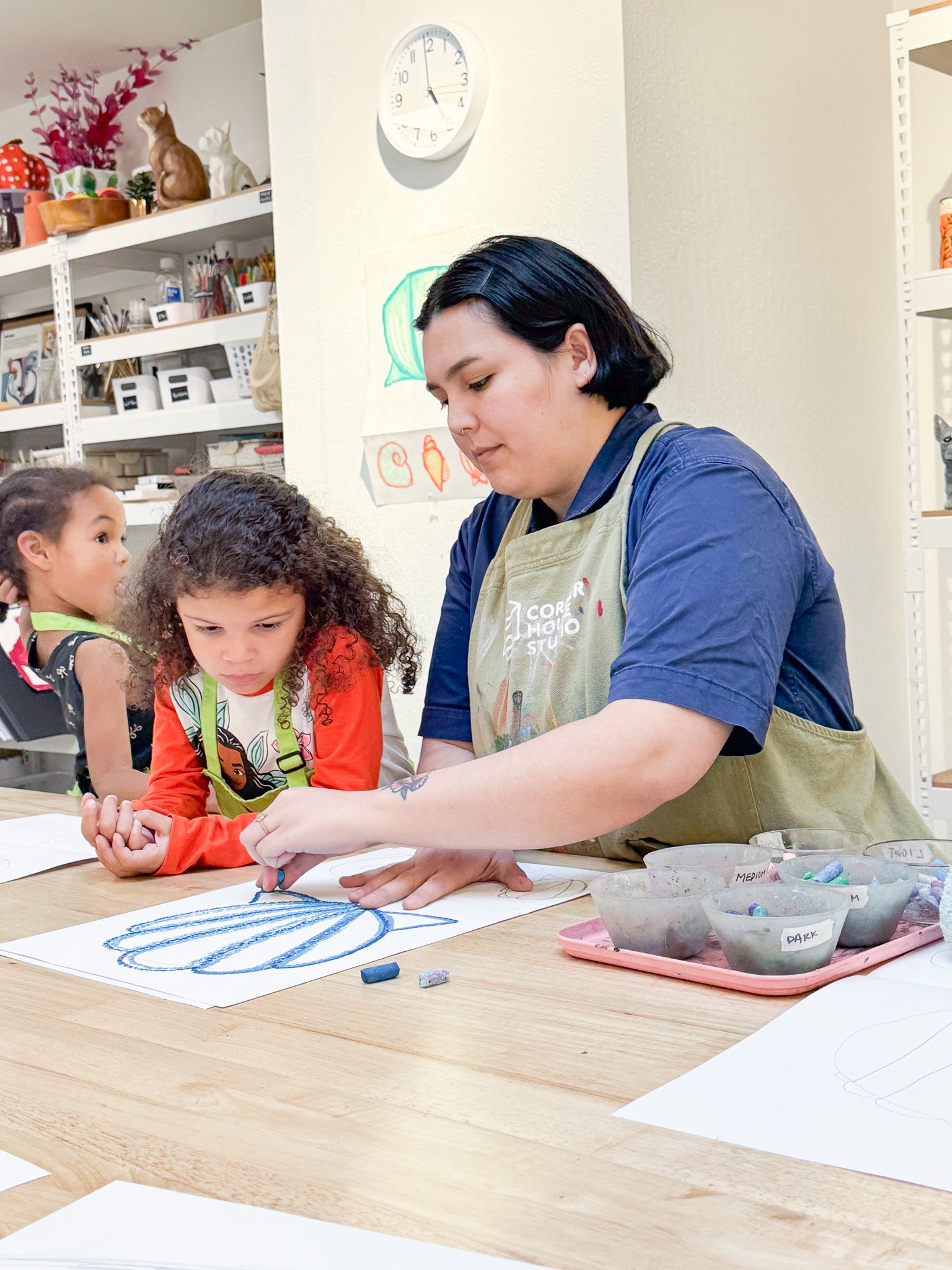 An adult woman and two young girls working on a craft project together at a table in an art classroom.