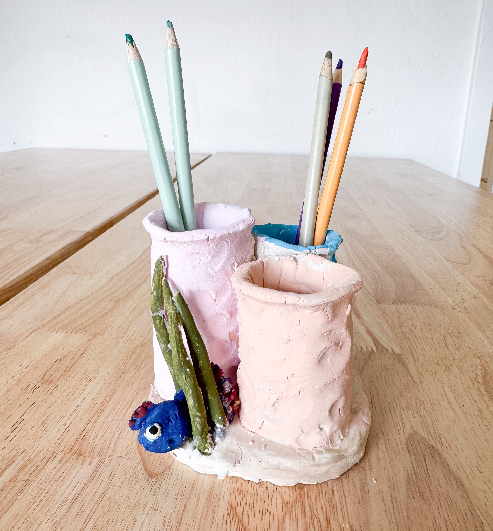 Handmade ceramic pencil holder with three compartments, decorated with a blue and a green caterpillar, containing pastel-colored pencils, placed on a wooden surface.