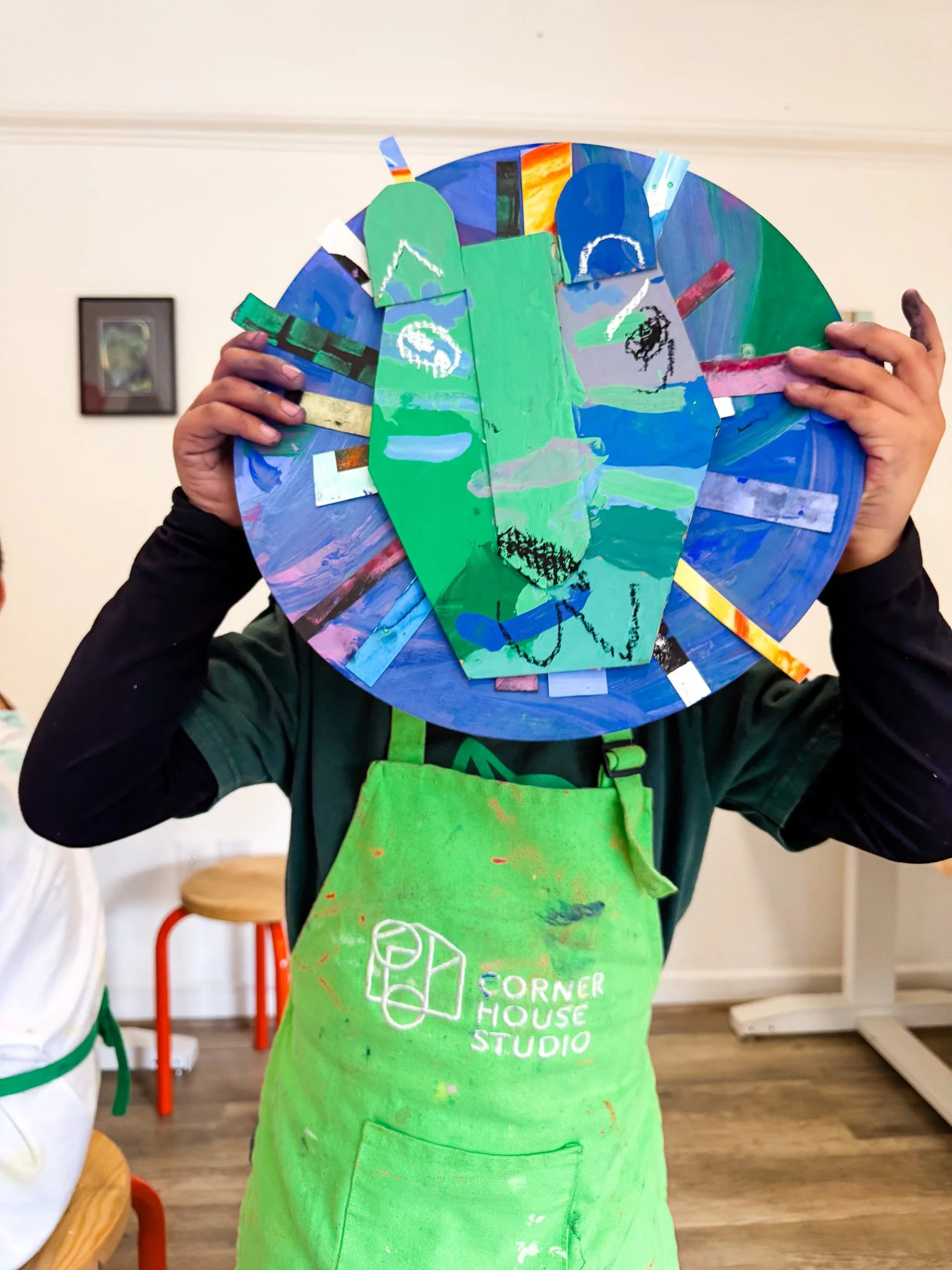 Child wearing a green apron holding a colorful, circular abstract painting in front of their face.