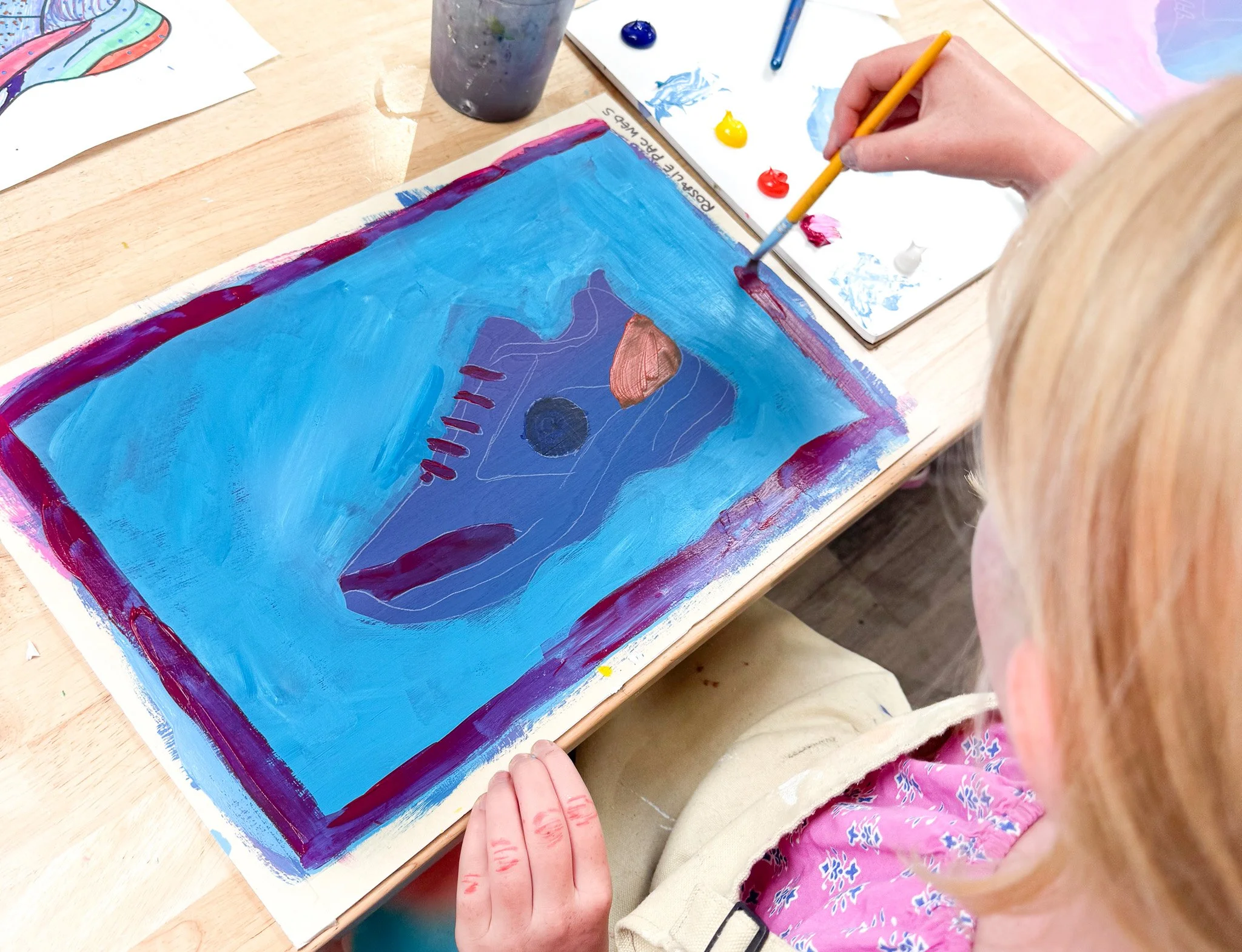 A person with blonde hair creates a blue abstract painting on paper with acrylic paints, using a small brush. The painting features various shapes and colors, including a black circle, orange, red, and purple accents, on a wooden table with a palette