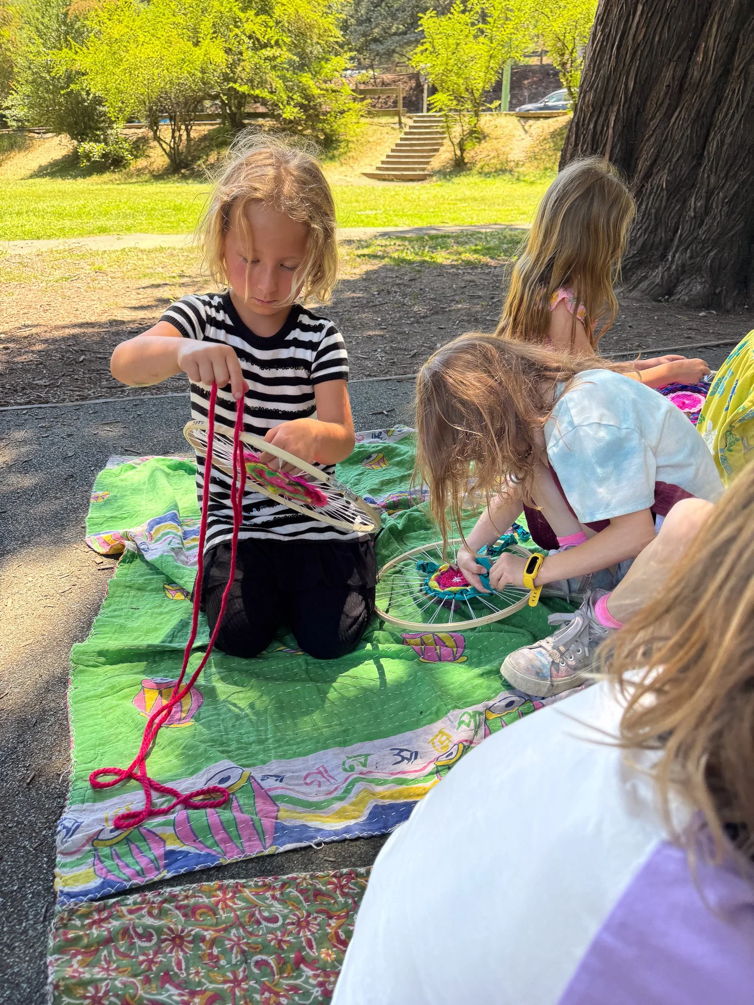 Three young girls are sitting outdoors on a green blanket with a colorful pattern, engaged in craft activities involving yarn and hoops, under a large tree with lush green leaves, on a sunny day.