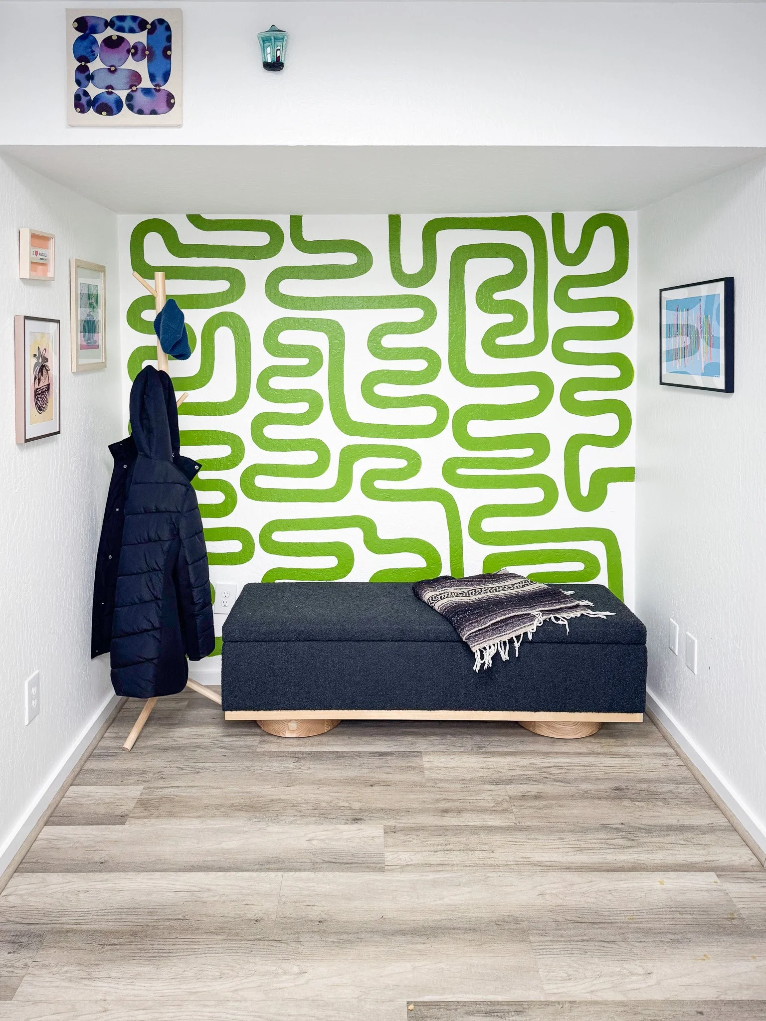 Small room with a green maze-patterned wall, a black bench with a throw blanket, artwork on the white walls, a coat rack with a jacket and hat, and ceiling decorations including a lantern.