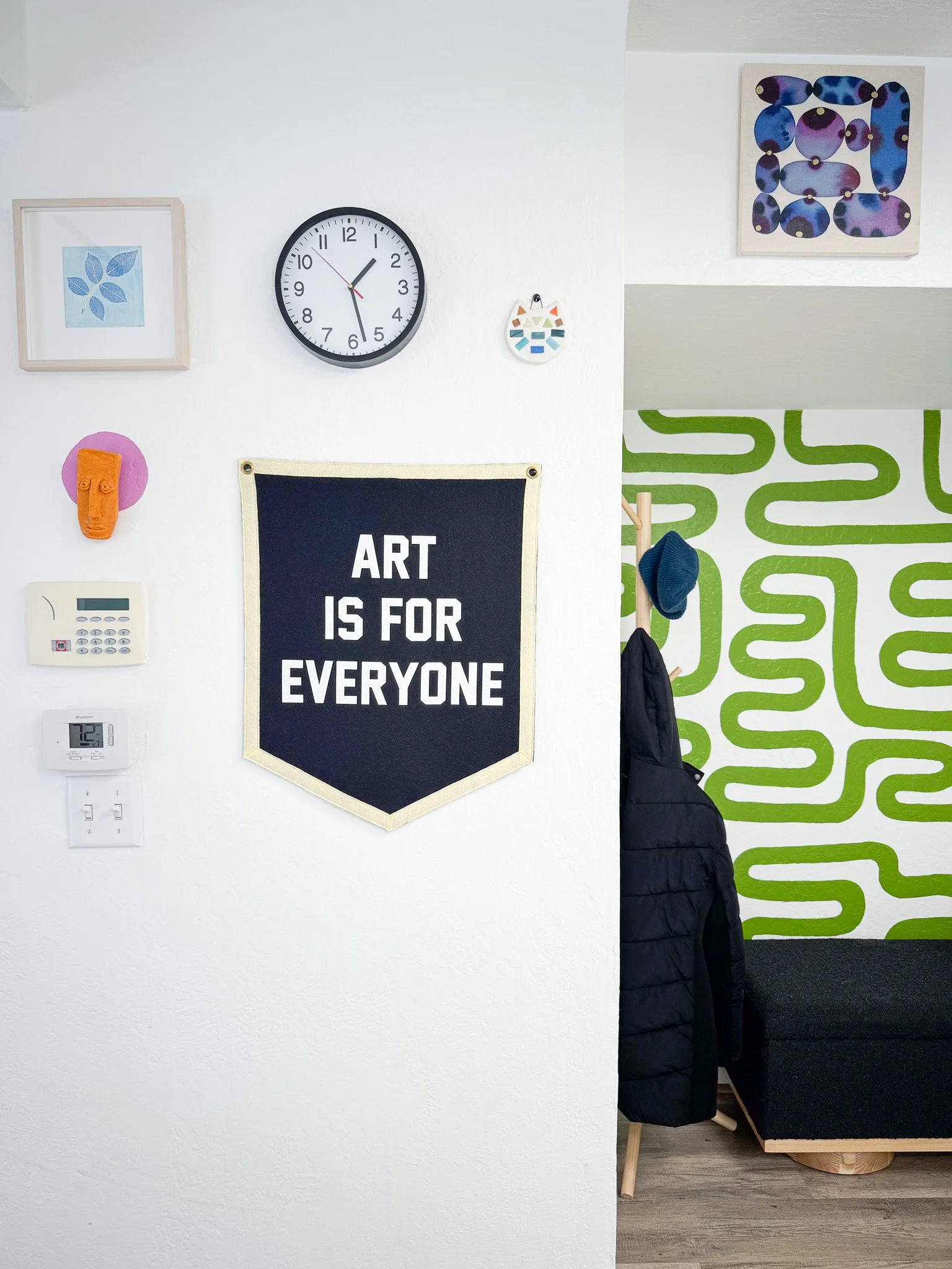 Interior wall with artwork, a clock, a decorative mask, a sign that says "Art is for Everyone," and some controls, with a partial view of a coat rack and a black couch in a colorful room.