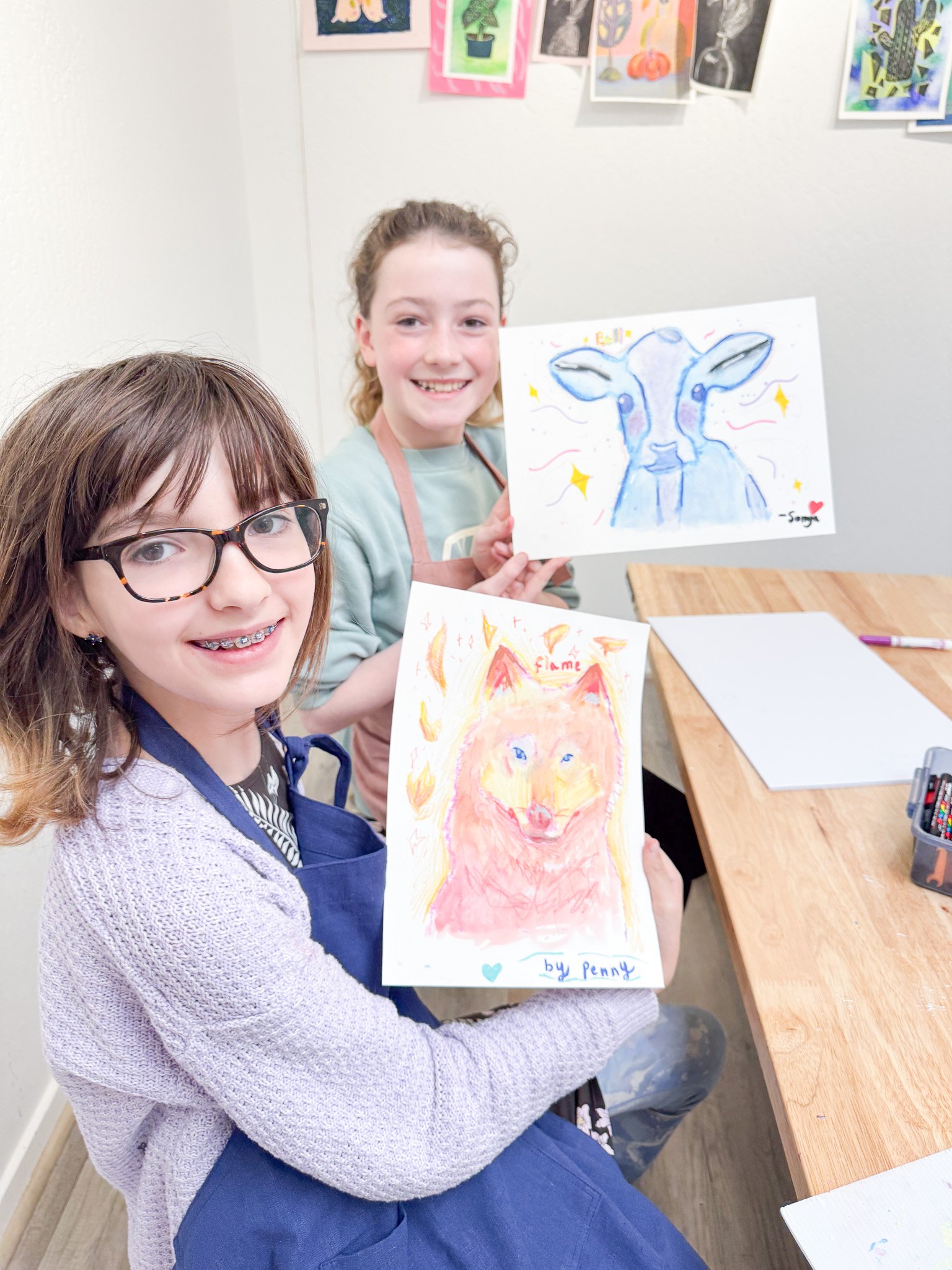 Two young girls are proudly displaying their watercolor animal paintings in an art classroom. The girl in the foreground has glasses and braces and holds a painting of a fox. The girl behind has curly hair and holds a painting of a blue and white goat. Artistic artwork and colorful art on the wall are visible.
