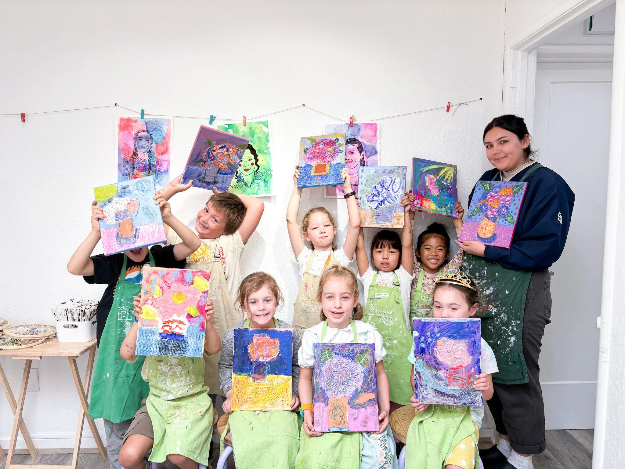 Group of children holding up their colorful artwork while an adult teaches in an art classroom.