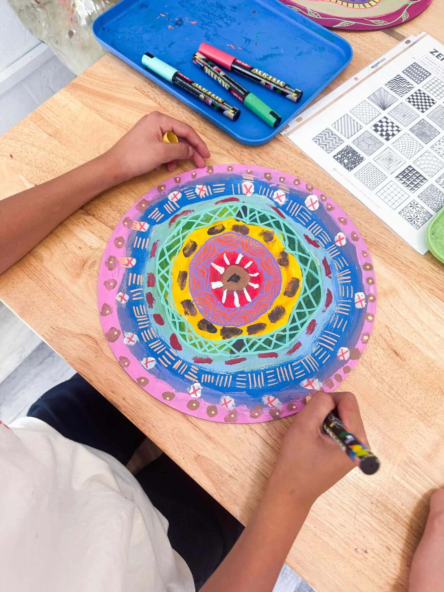 Child creating a colorful, circular mandala on paper with markers, on a wooden table, with art supplies around.